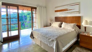 A bedroom with a large bed, a wooden headboard, a nightstand, a lamp, and a view of greenery through a sliding glass door. - Pousada Golf Village (Petrópolis)
