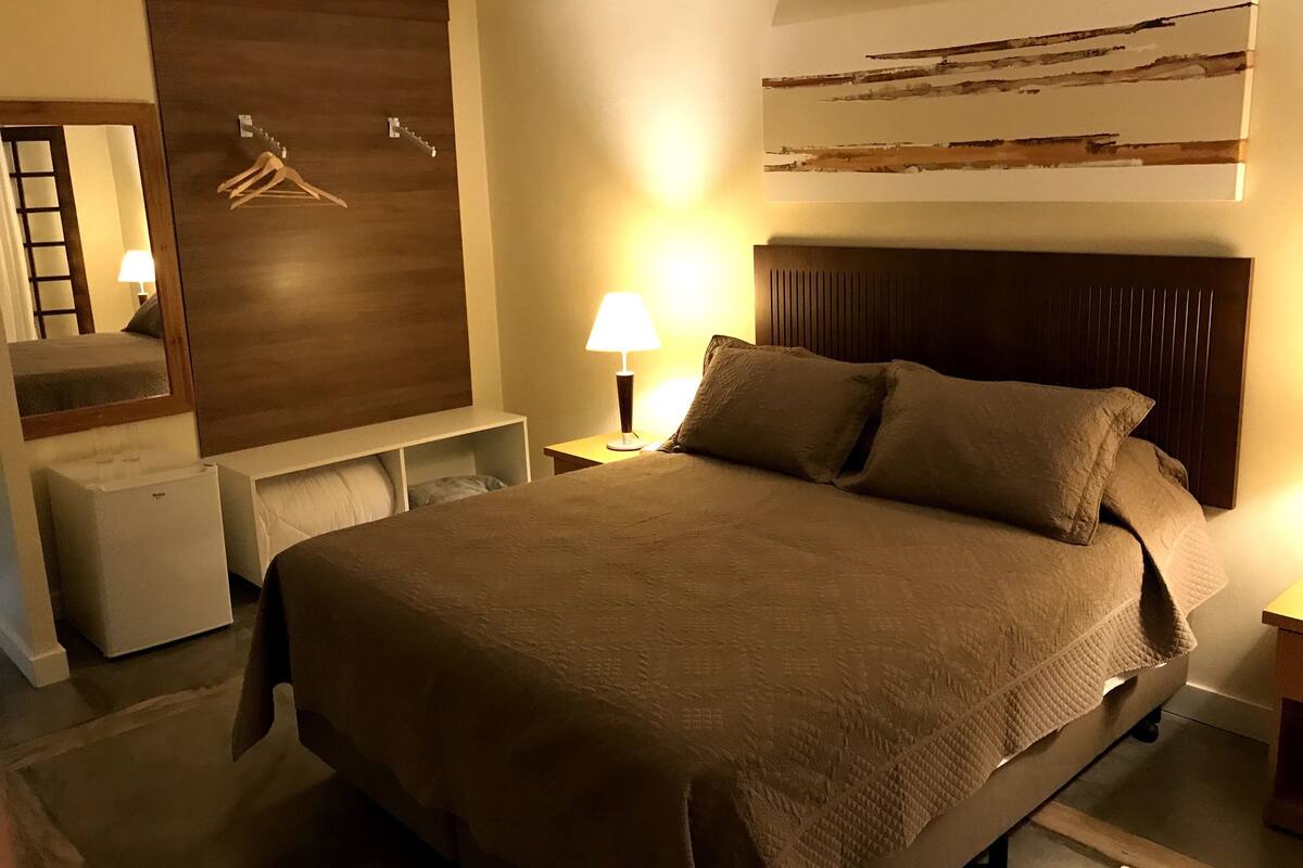 A neatly made bed with a headboard, a bedside lamp, a small table, a mirror, and a wall-mounted artwork.