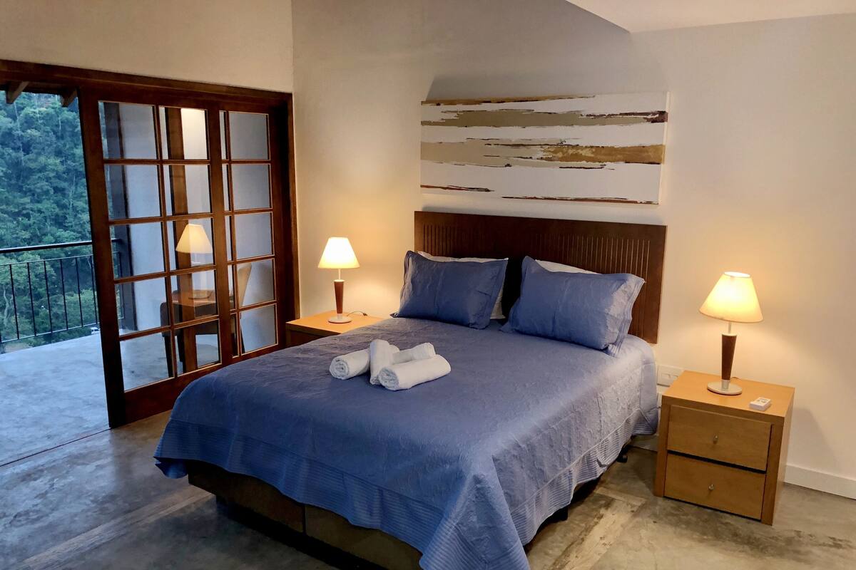 A bedroom with a bed, two bedside tables, a nightstand, a large window, and a view of greenery outside.