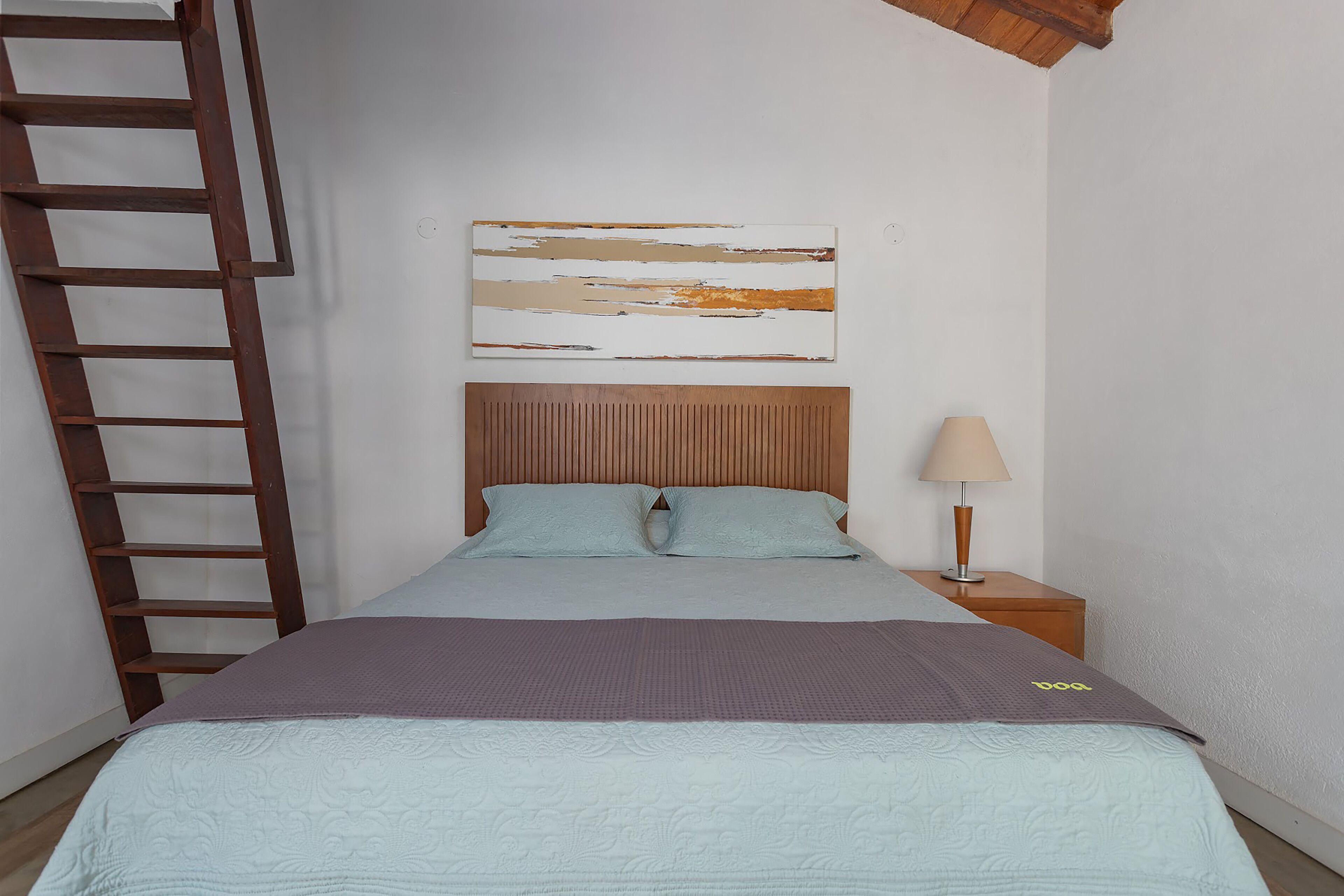 A bedroom with a bed, a wooden headboard, a nightstand with a lamp, and a ladder against the wall.