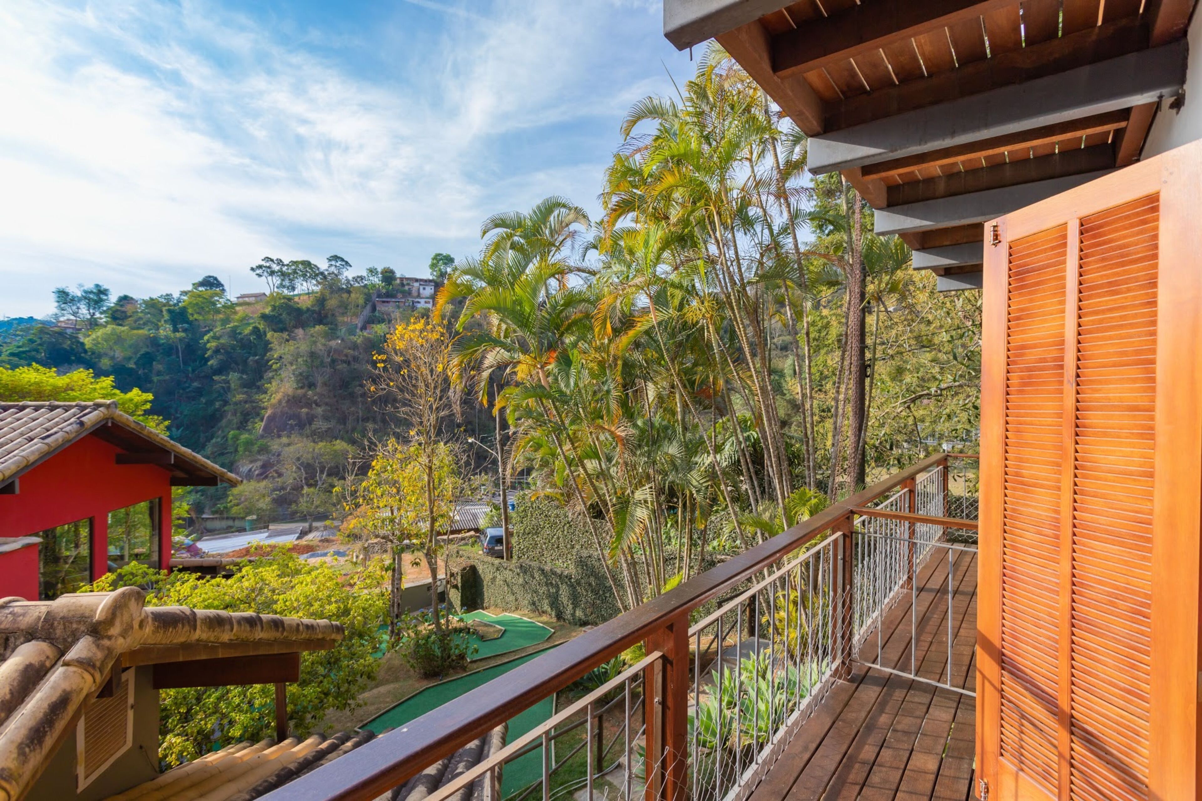 A balcony with a view of a lush garden, a wooden deck, and a red building.