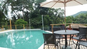 A pool area with a wooden deck, metal chairs, and a table with an umbrella. - Pousada Golf Village (Petrópolis)