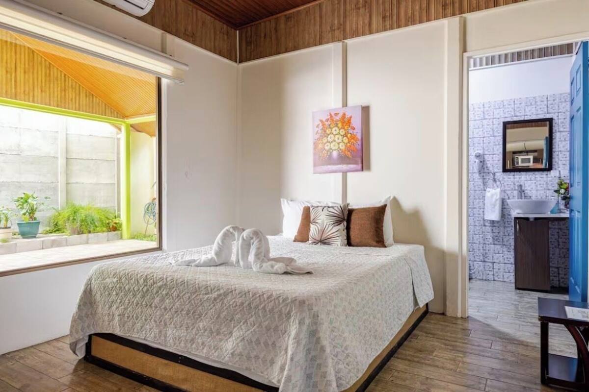 A hotel room with a bed, a window with plants, a painting, and a bathroom visible through an open door.