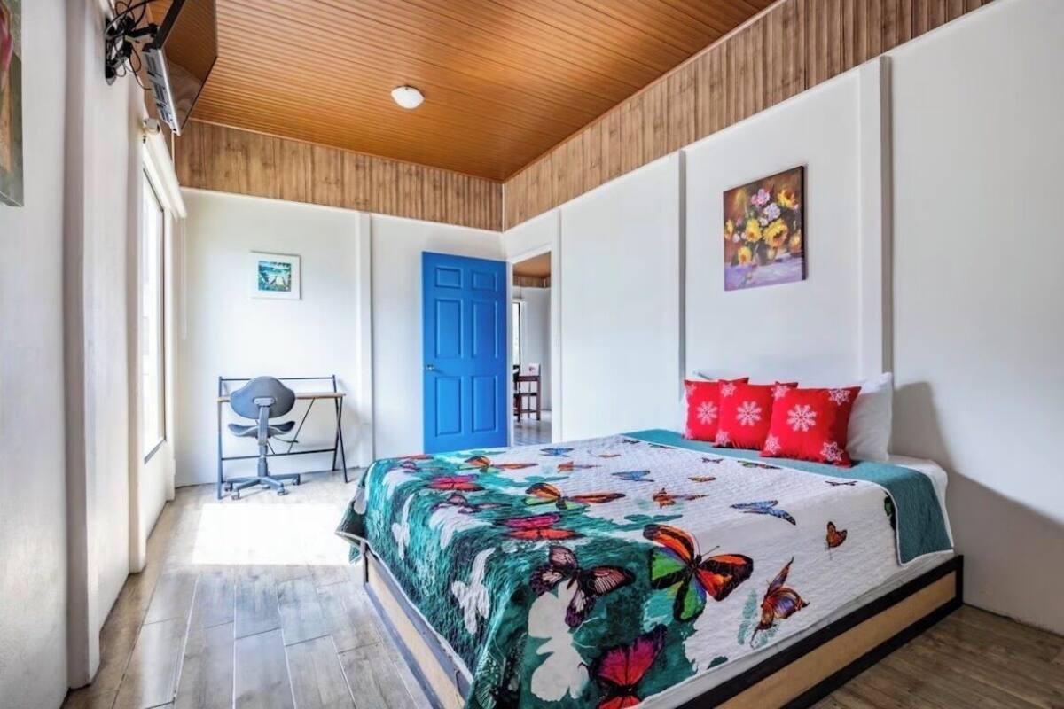 A bedroom with a wooden ceiling, a bed with a colorful butterfly quilt, a blue door, and a small desk with a chair.
