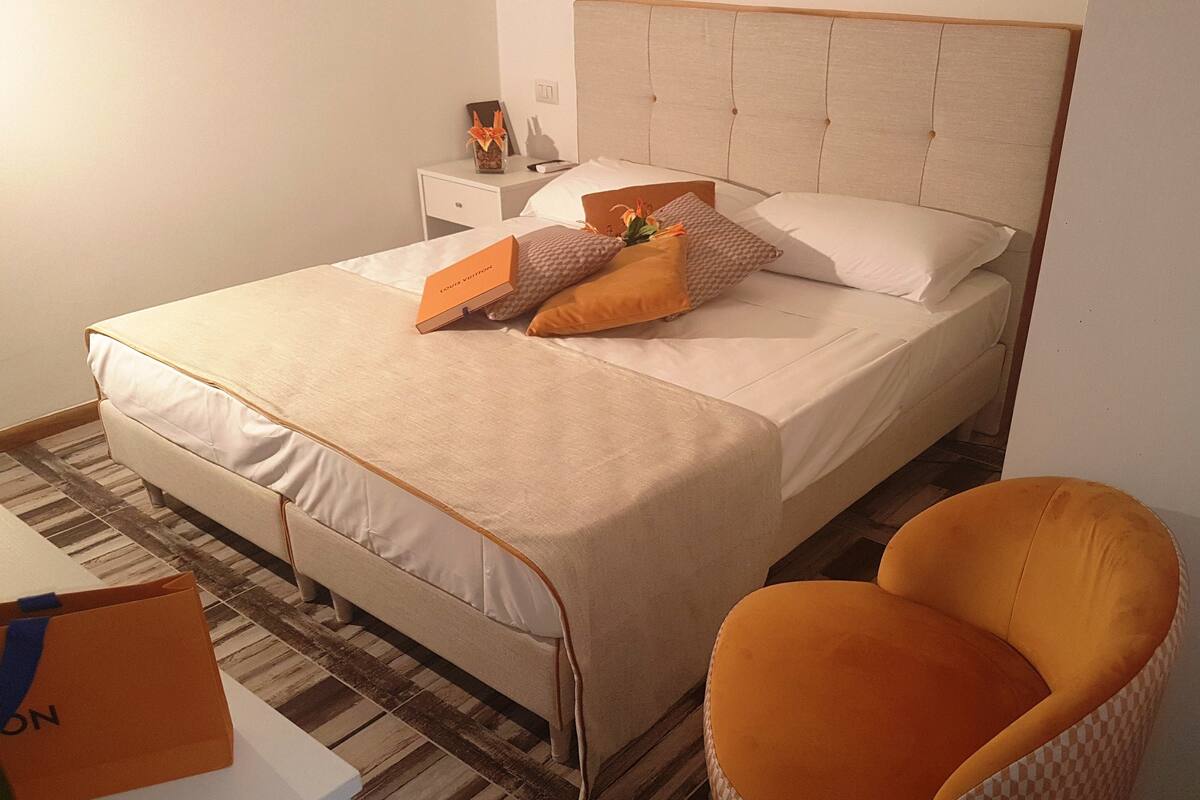 Cama bem arrumada com cabeceira, mesa de cabeceira com abajur e uma cadeira laranja.