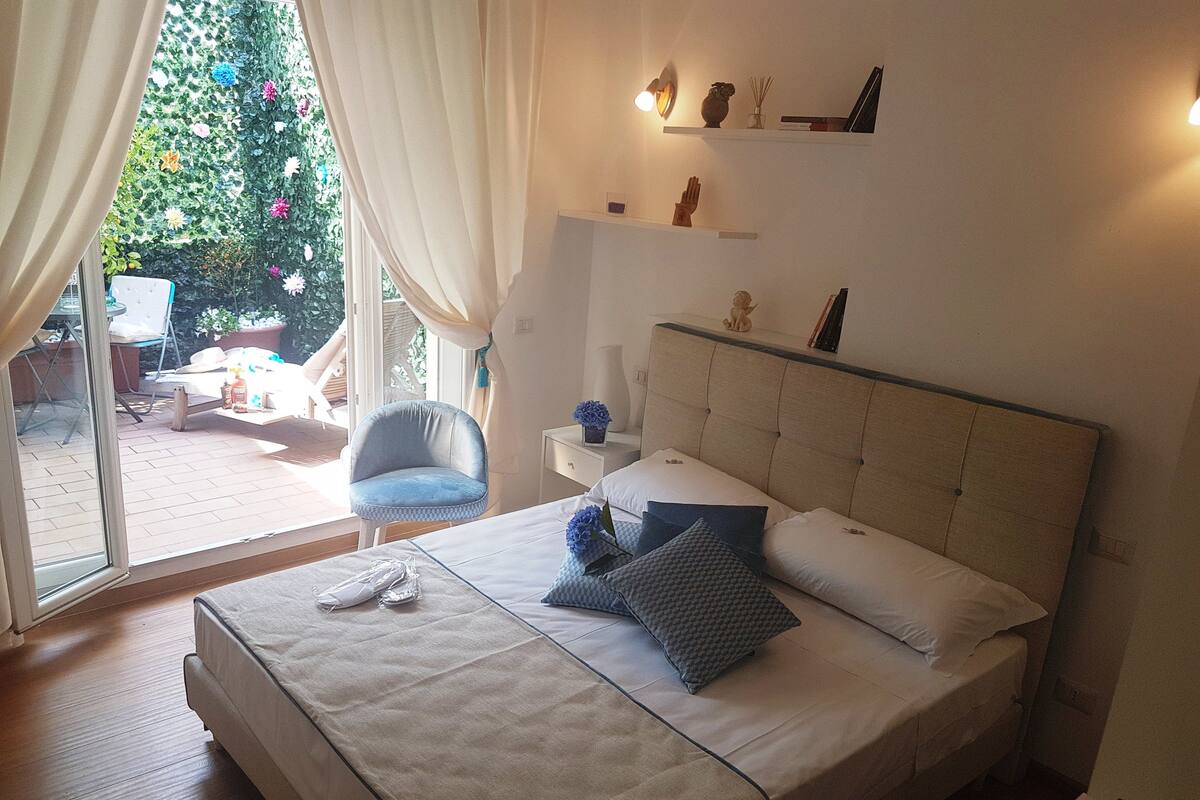 Quarto com cama grande, uma cadeira, mesa de cabeceira e janela com cortinas.