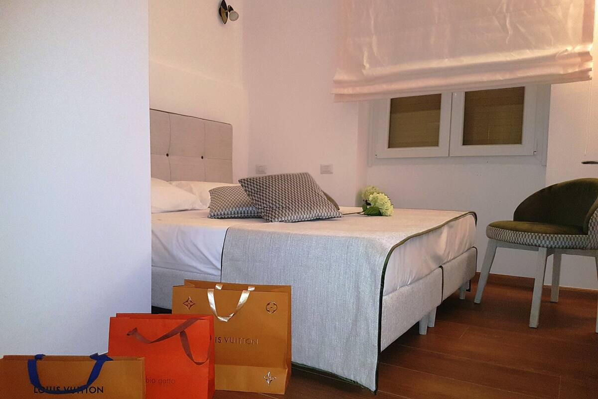 Quarto de hotel com uma cama, uma cadeira e duas sacolas de compras Louis Vuitton no chão.