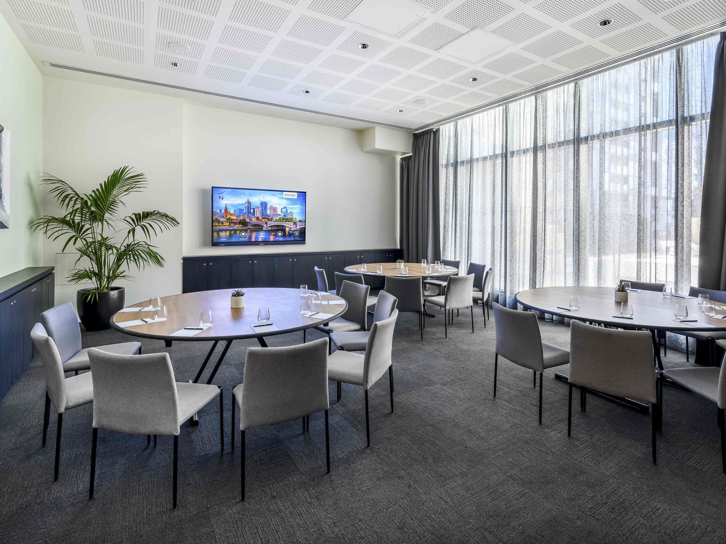 Un salón de conferencias con mesas redondas, sillas, una planta grande en maceta, un televisor de pantalla plana y ventanas amplias con cortinas.