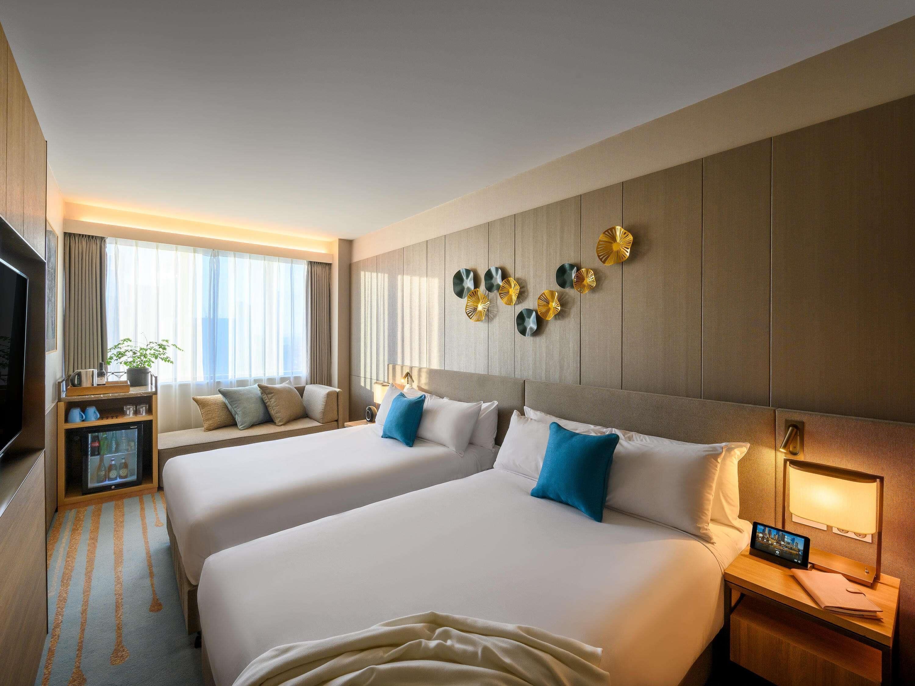 A hotel room with two beds, a TV, a bedside table with a lamp, and a wall-mounted clock.