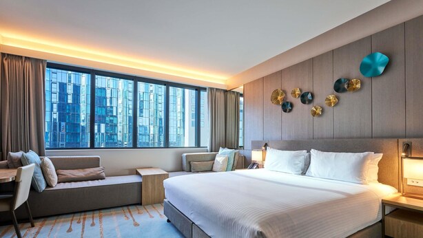Une chambre d’hôtel moderne dotée d’un grand lit, d’un coin salon, d’un bureau et offrant une vue sur le paysage urbain grâce à de grandes fenêtres.