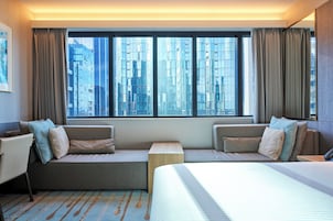 A modern hotel room with a large window, a sofa bed, a bedside table, and a bed.
