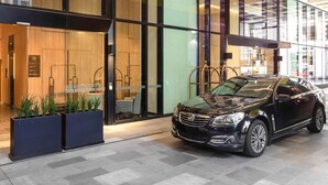L’entrée d’un hôtel moderne, avec une voiture noire garée devant, des portes en verre et un ascenseur bien visible.