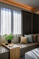Ein modernes Hotelzimmer mit einer Couch, einem kleinen Tisch mit Pflanze und einem Fenster mit durchsichtigen Vorhängen.