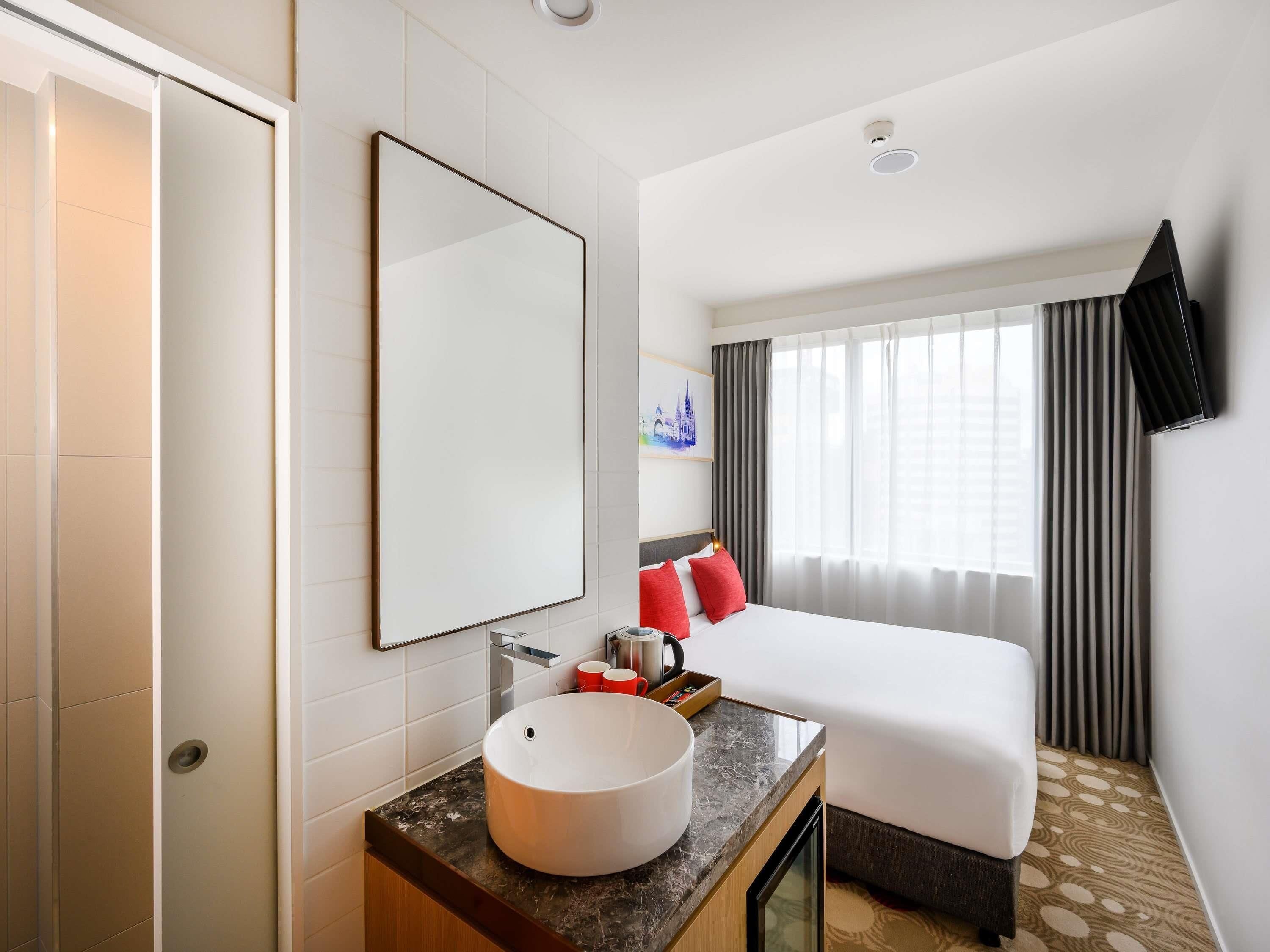 Une chambre d’hôtel avec un lit, un grand miroir, un lavabo rond et une fenêtre avec des rideaux.