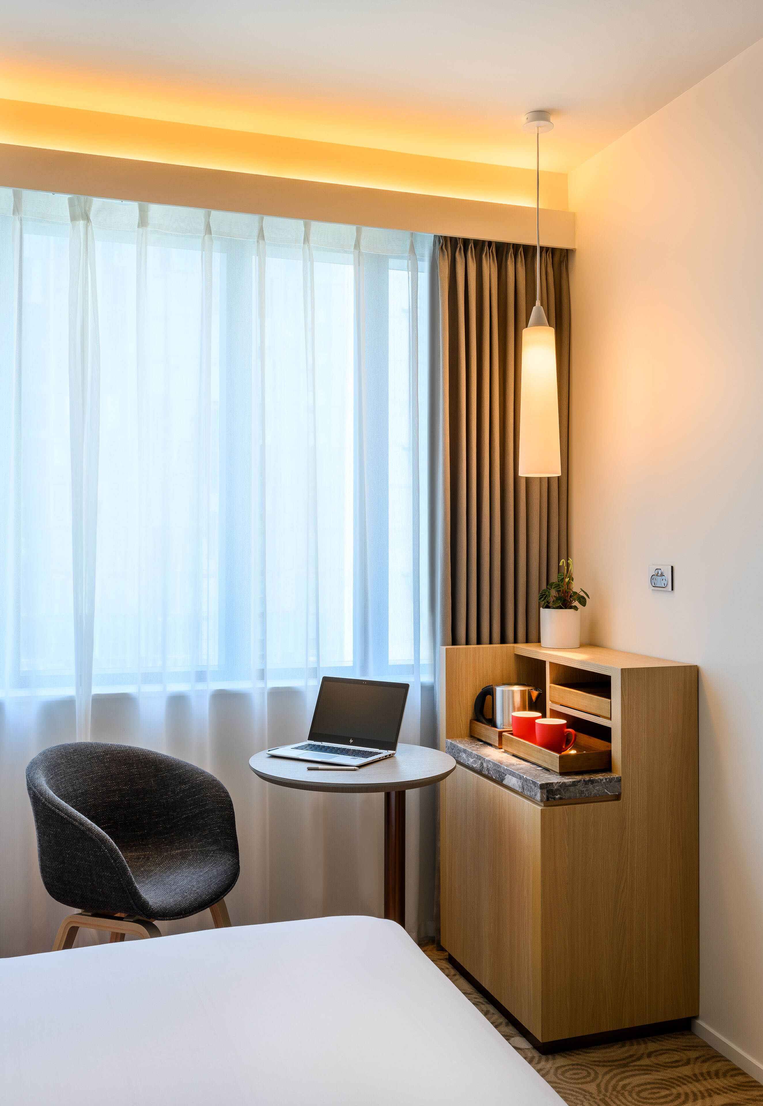 A hotel room with a bed, a desk with a laptop, a chair, a small table, and a built-in wooden cabinet with a plant and red mugs.