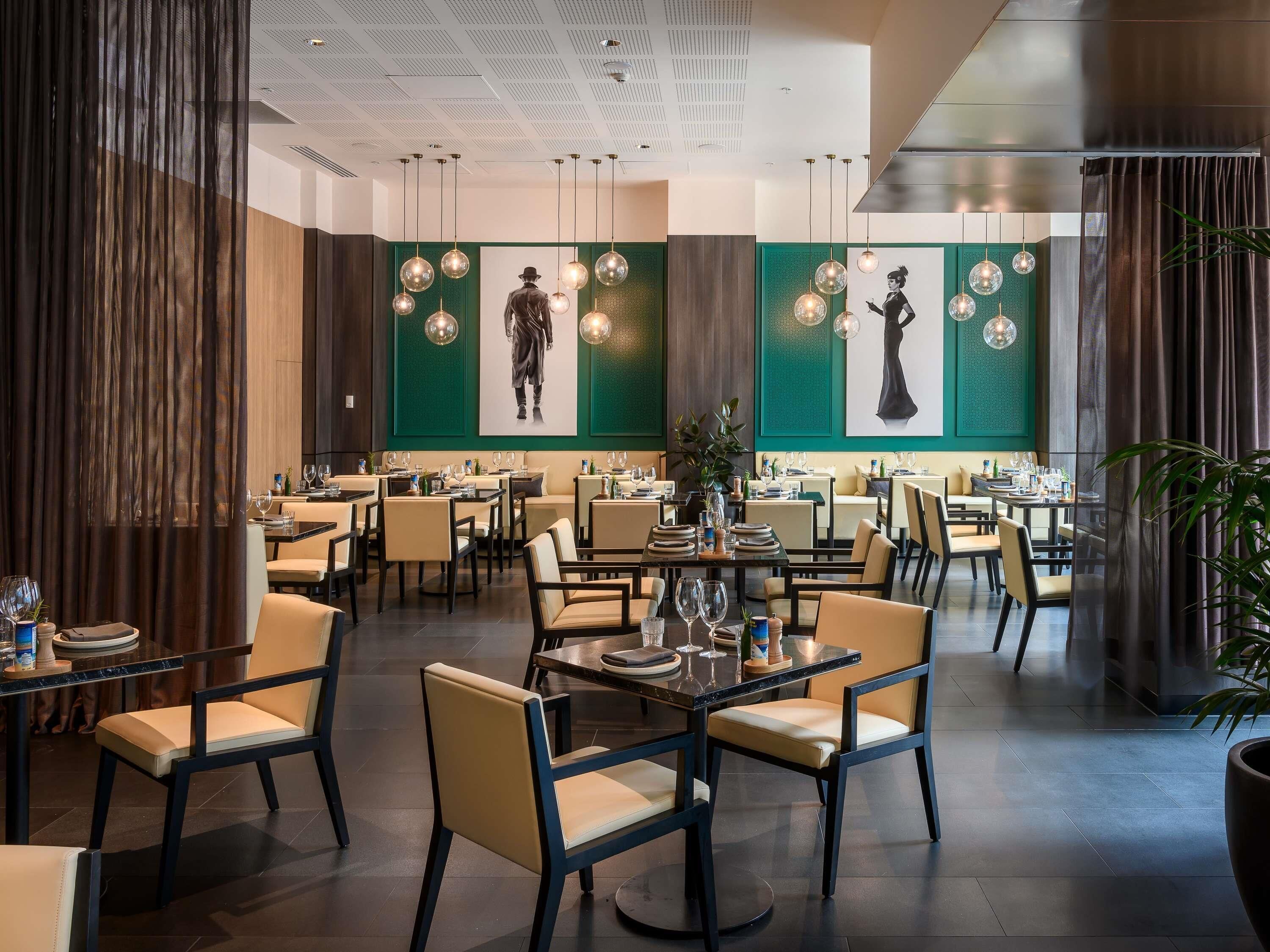 Un restaurante moderno con varias mesas y sillas para comer, paredes verdes con obras de arte, iluminación colgante y un techo con diseño de cuadrícula.