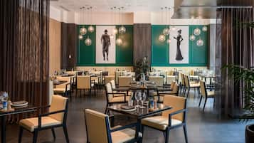 Un restaurante moderno con varias mesas y sillas para comer, paredes verdes con obras de arte, iluminación colgante y un techo con diseño de cuadrícula.