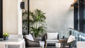 Un área de estar exterior moderna con sillas de mimbre y una mesa blanca, rodeada de plantas en macetas y un gran árbol.
