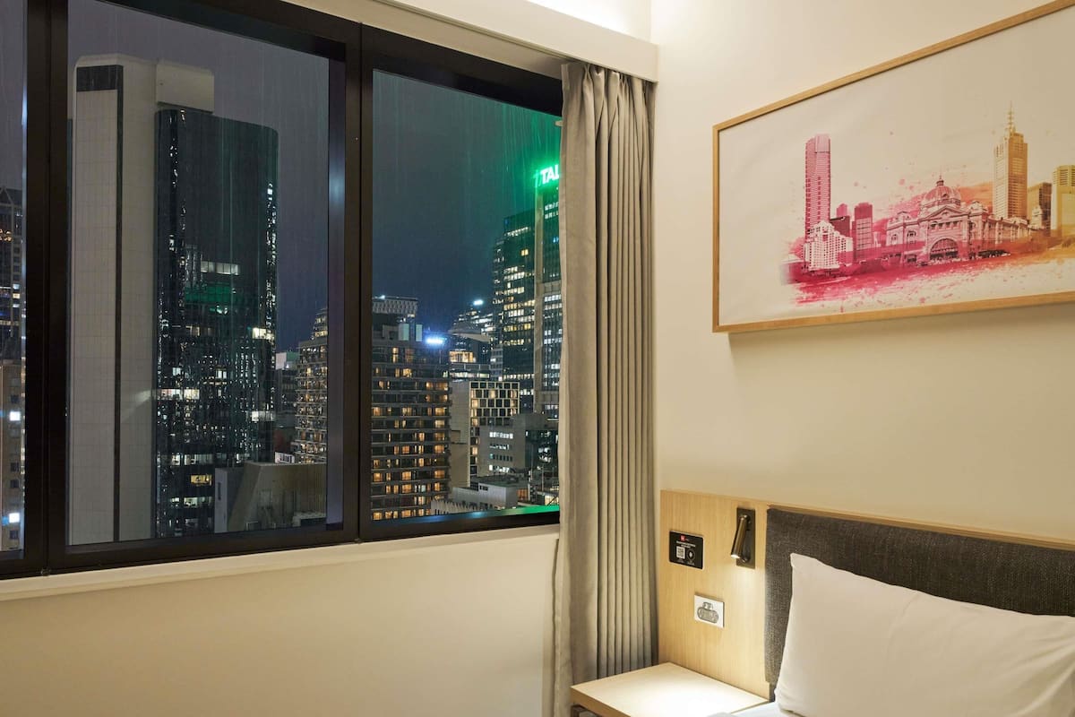 A hotel room with a cityscape view through a window, a bed, a bedside table, a nightstand, and a wall-mounted painting.