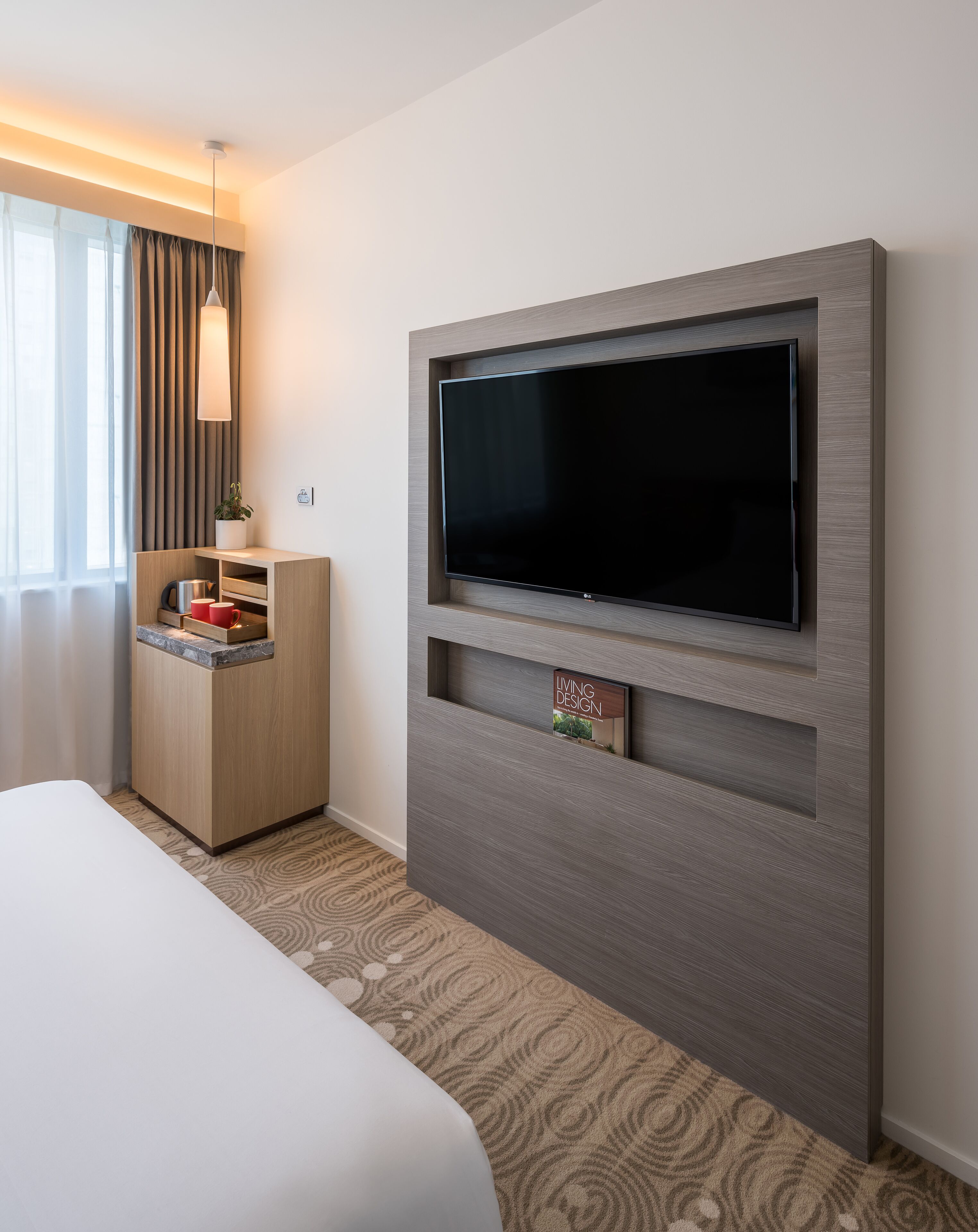 A hotel room with a large flat-screen TV mounted on a wooden wall, a small wooden cabinet with a red mug, and a patterned carpet.