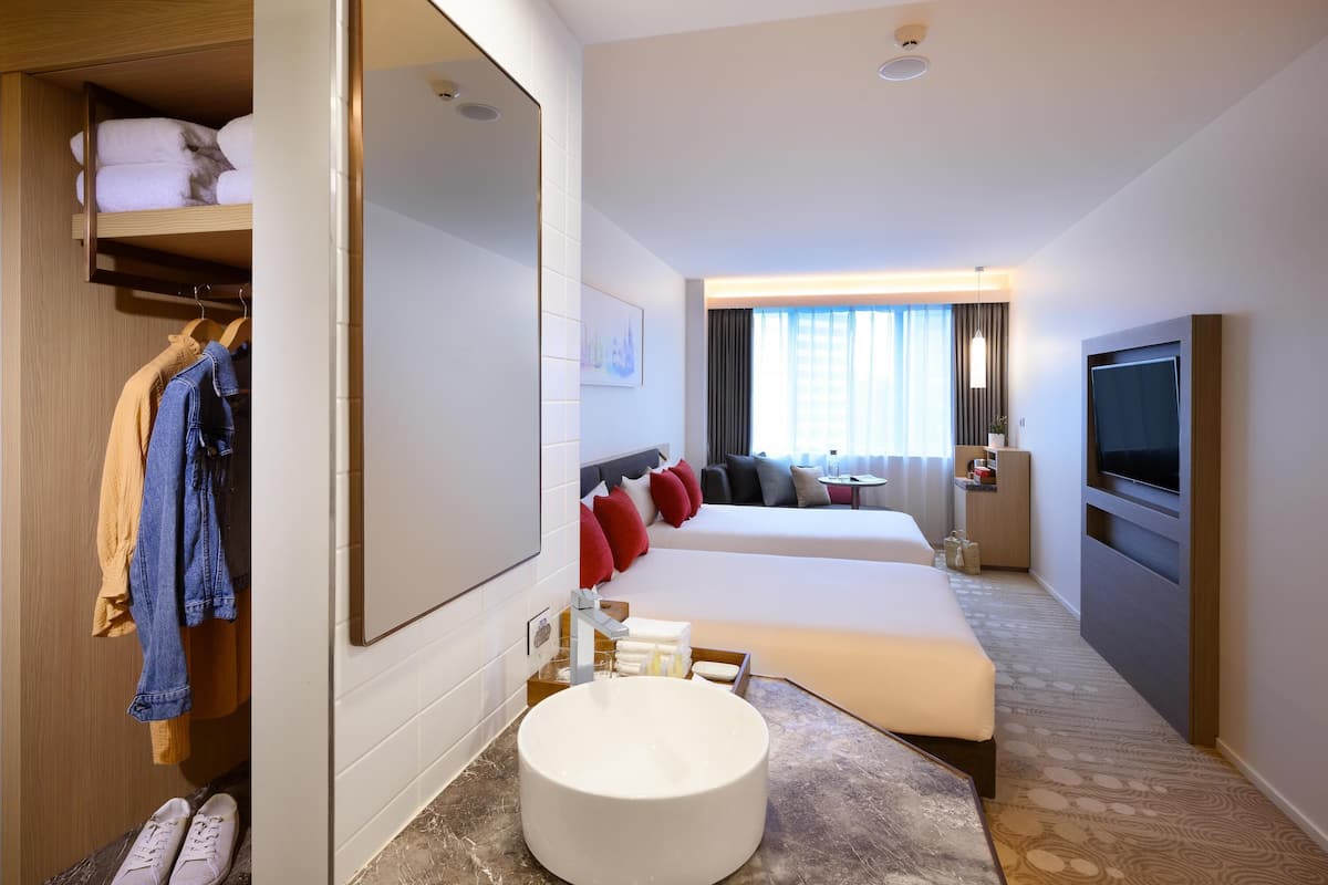 A hotel room with a large mirror, a round sink, a bed, a TV, and a wardrobe with hanging clothes.