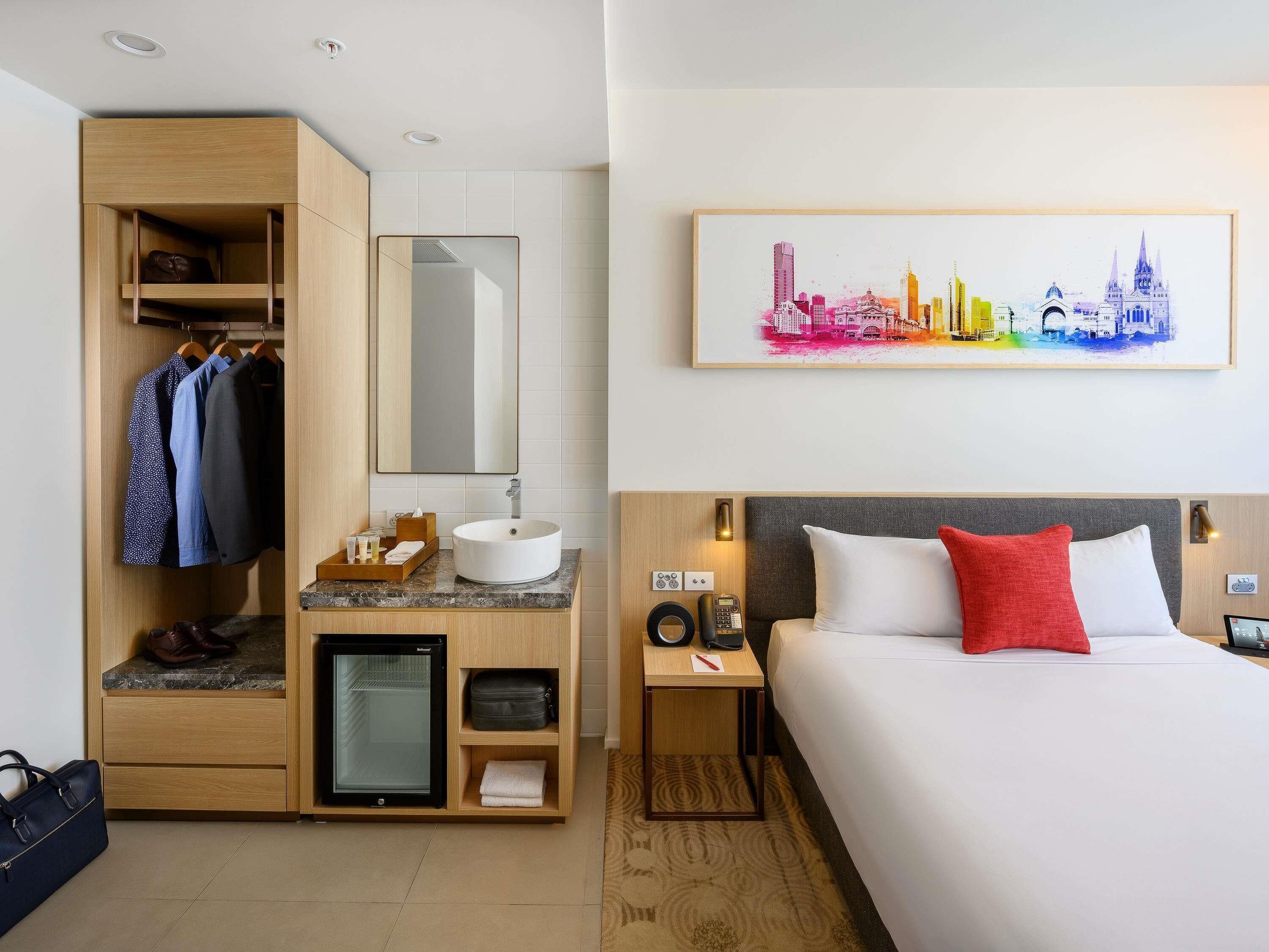 A hotel room with a bed, a desk with a sink, a minibar, a wardrobe, and a cityscape painting on the wall.