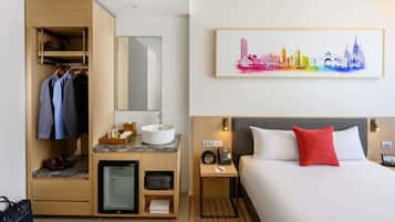 A hotel room with a bed, a desk with a sink, a minibar, a wardrobe, and a cityscape painting on the wall.