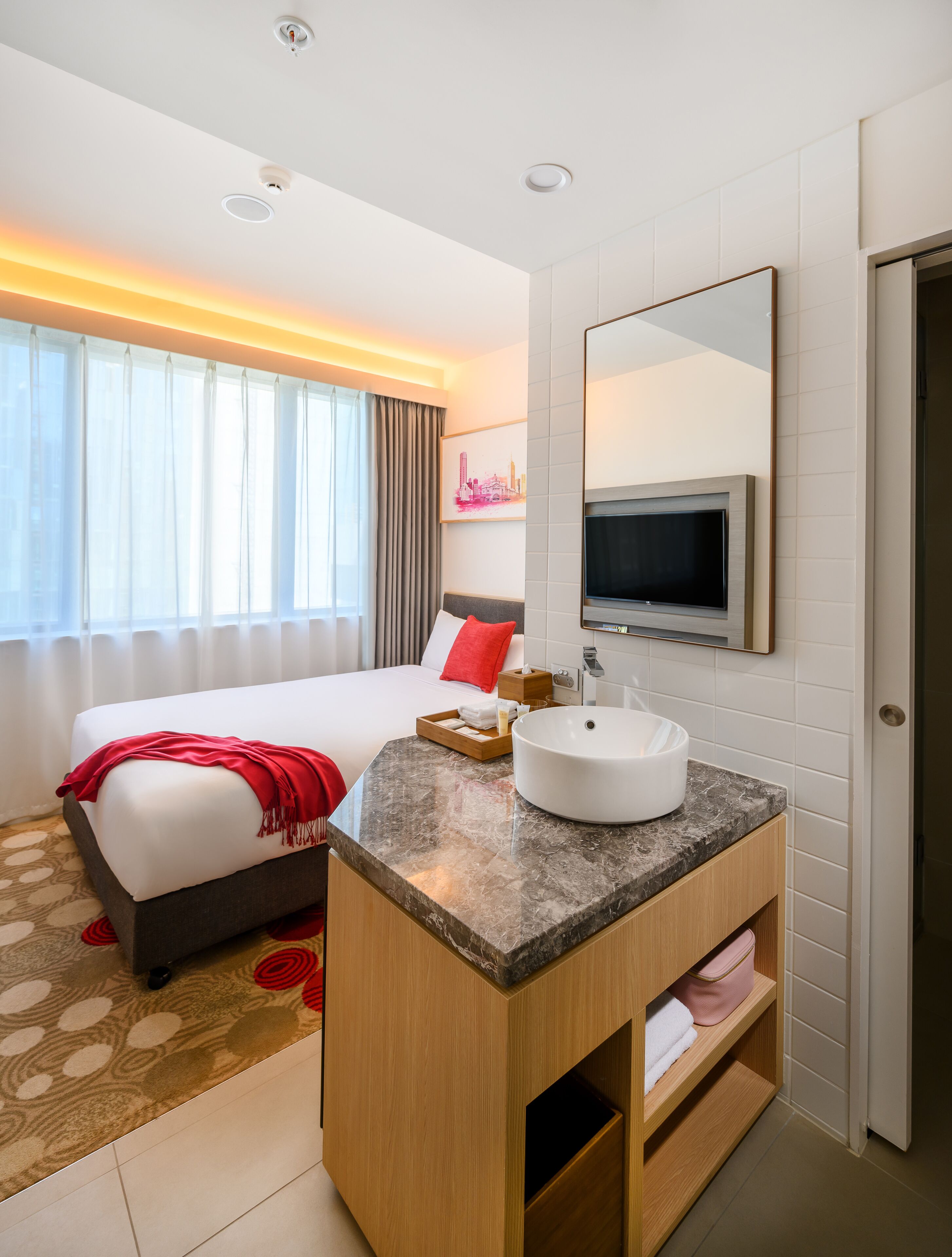 A hotel room with a bed, a bathroom vanity, a large mirror, and a TV mounted on the wall.