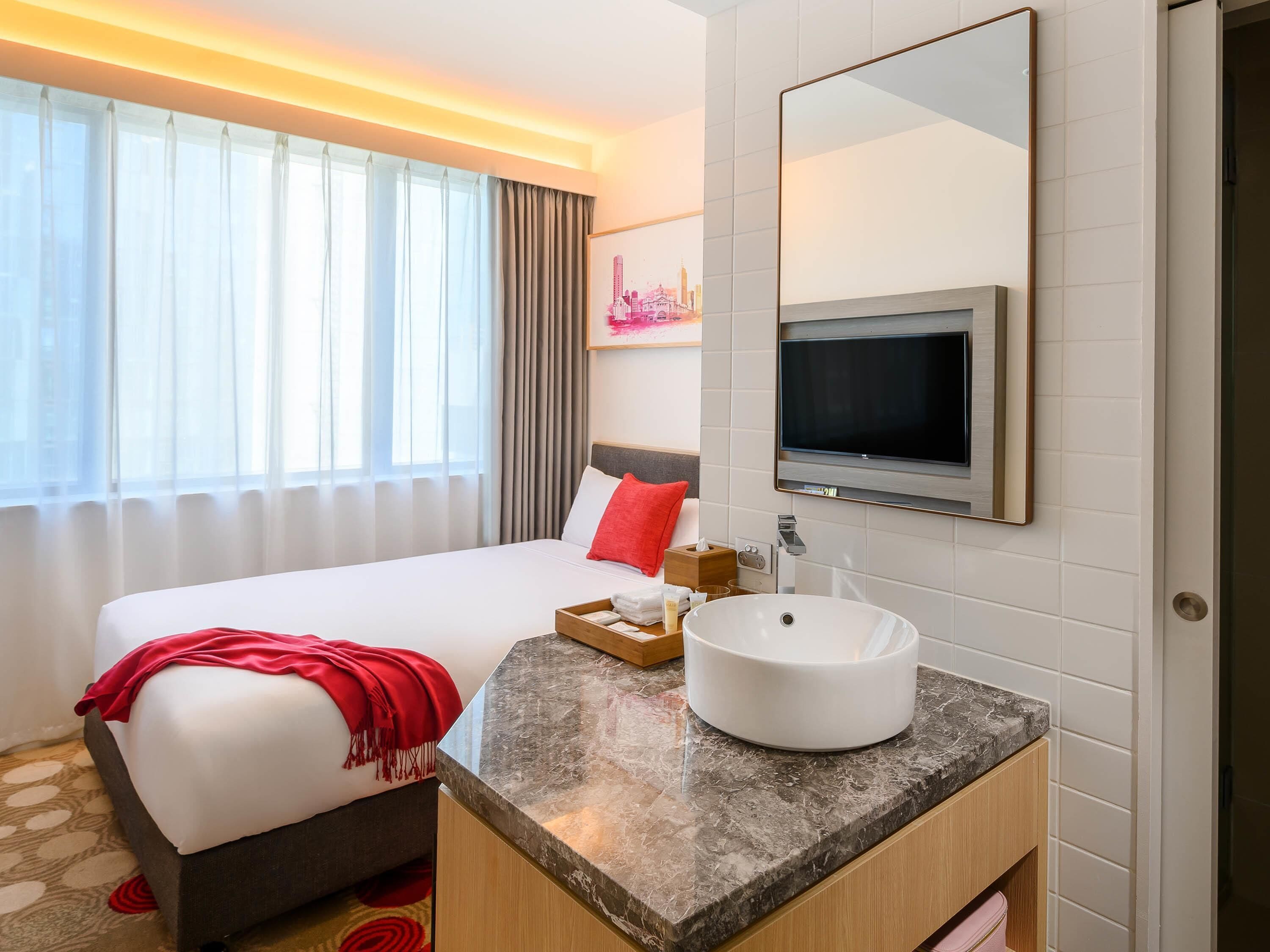 A hotel room with a bed, a bedside table with a white sink, a TV mounted on the wall, and a window with curtains.