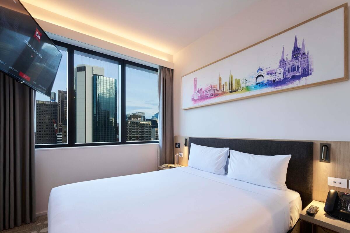 A hotel room with a large bed, a cityscape view, a flat-screen TV, and a painting of a city skyline.