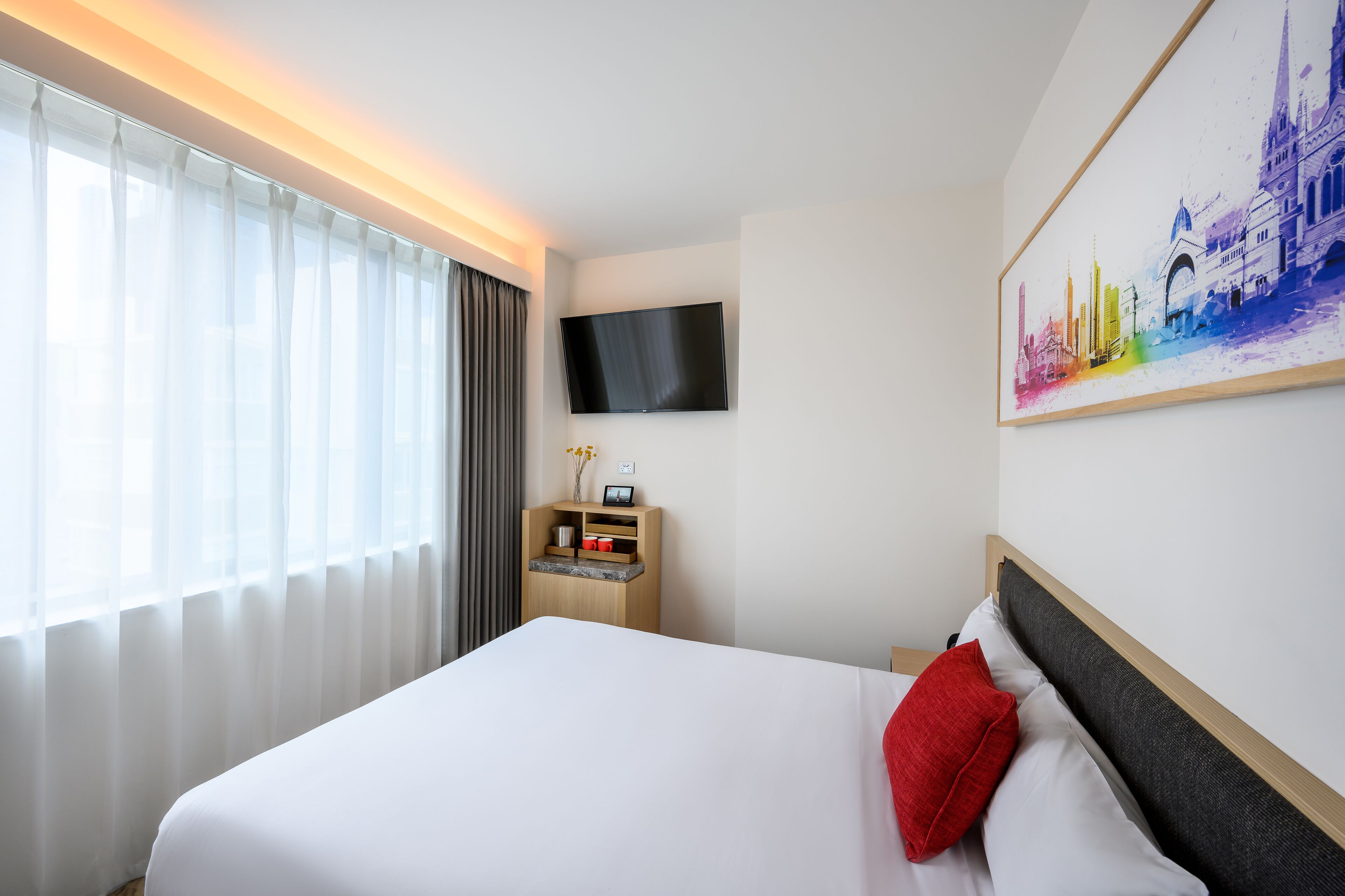 A hotel room with a bed, a TV mounted on the wall, a painting, and a small wooden cabinet with a vase and a picture frame.
