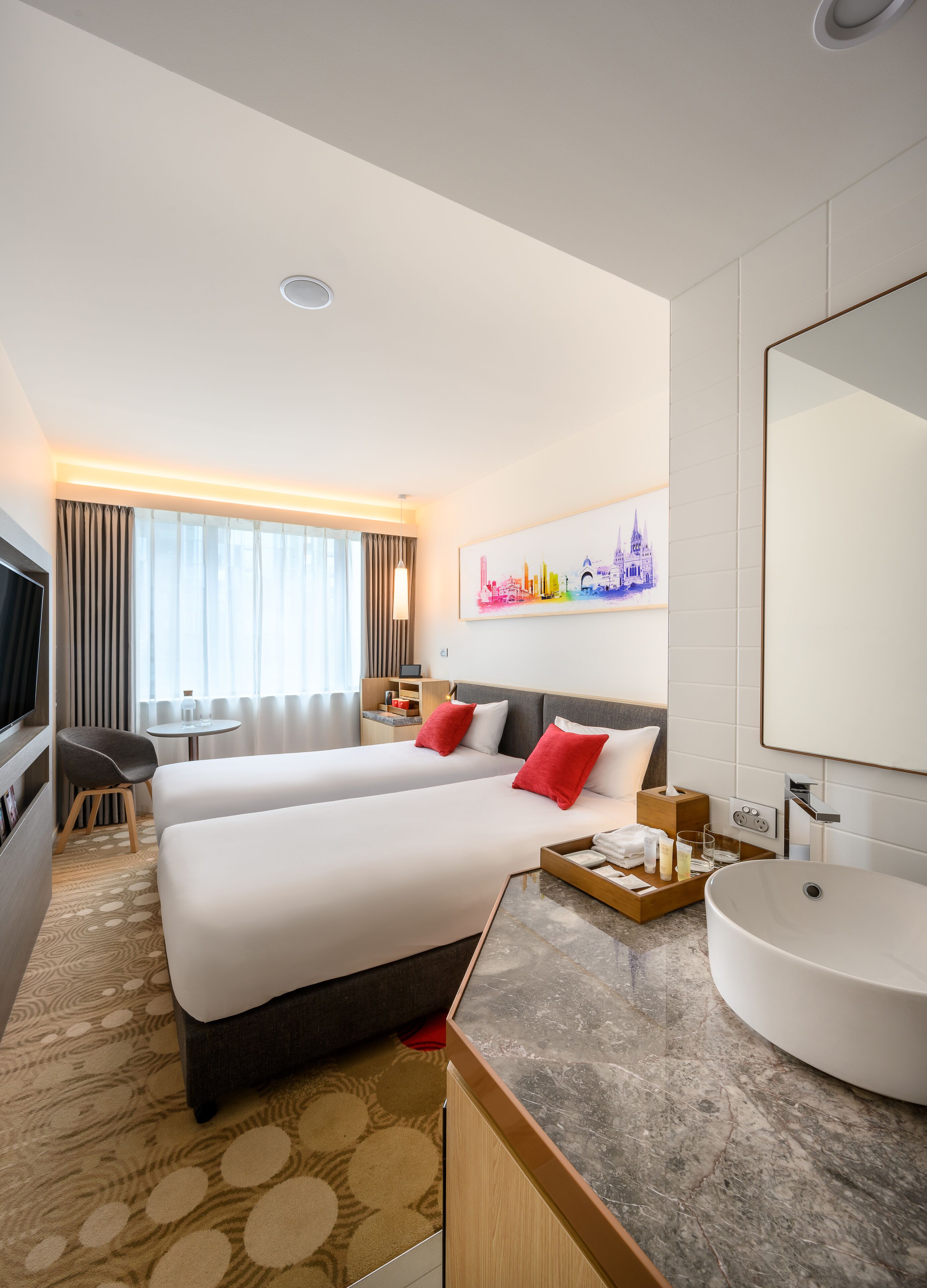 A hotel room with two beds, a round sink, a TV, a desk, and a cityscape artwork on the wall.