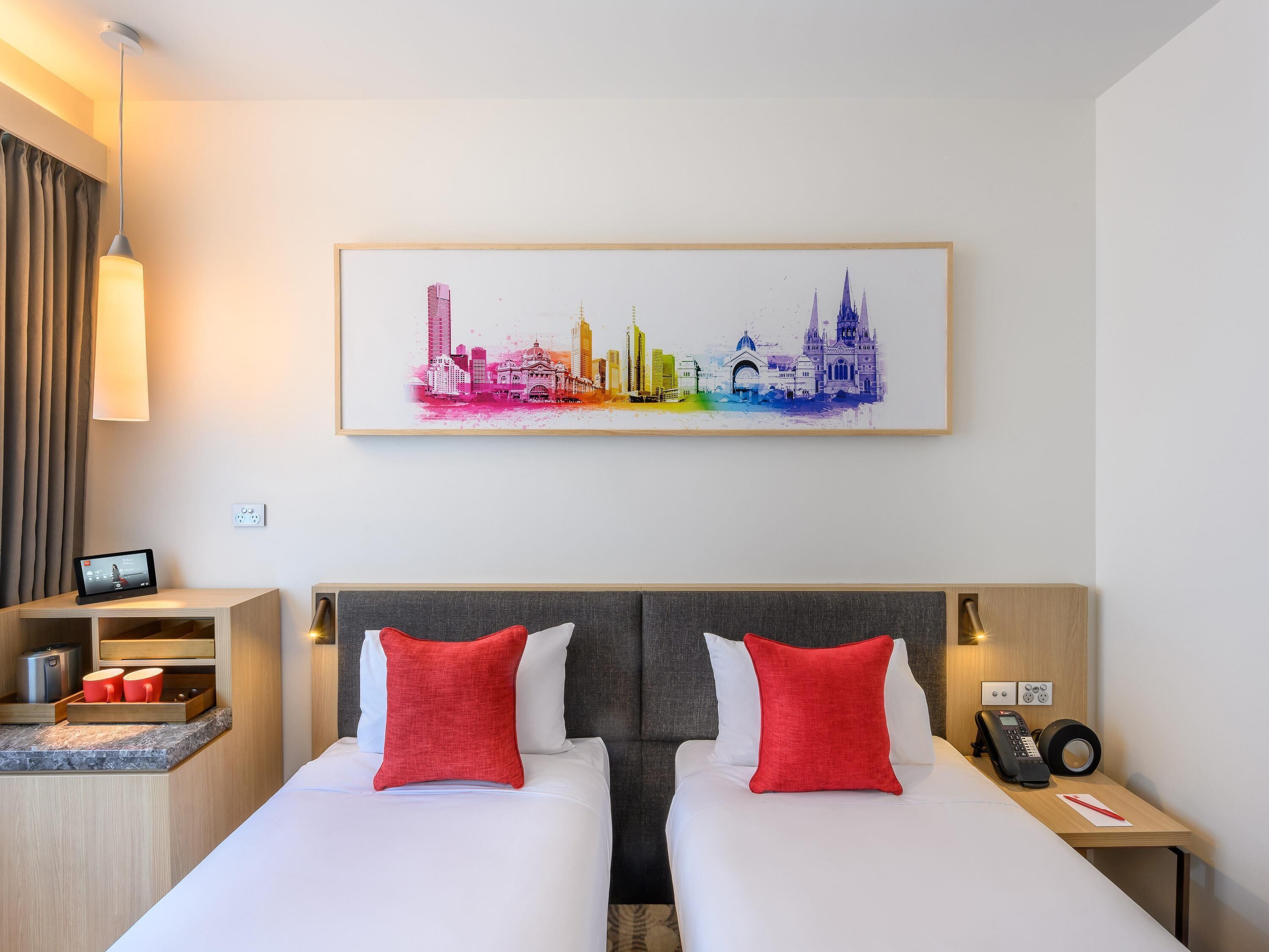 A hotel room with a bed, bedside tables, a small desk, a telephone, and a cityscape painting on the wall.