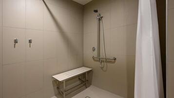 A small, modern shower room with a bench, a handheld showerhead, and a wall-mounted control panel.