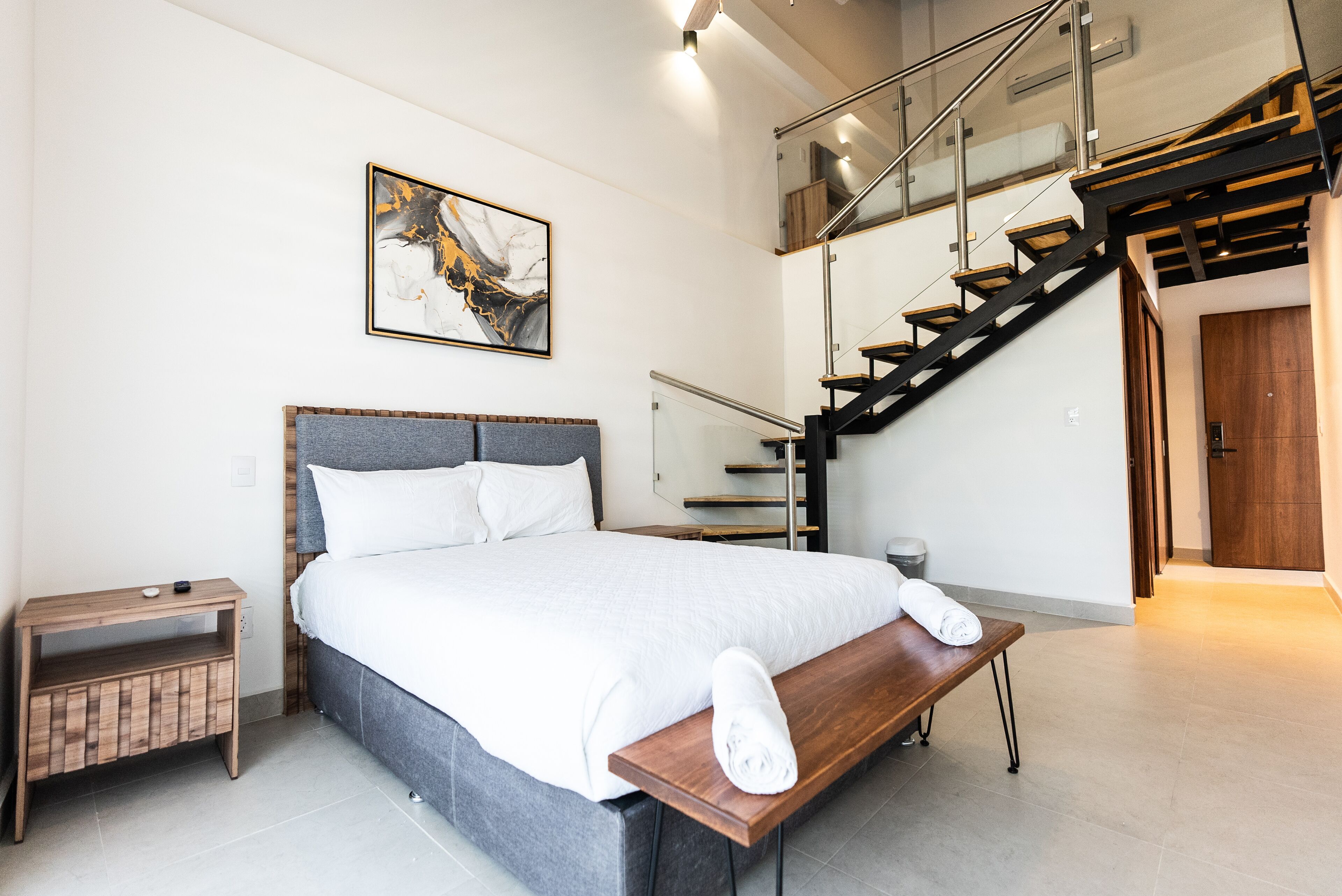 A modern hotel room with a large bed, a wooden bench, a nightstand, a painting, and a staircase.