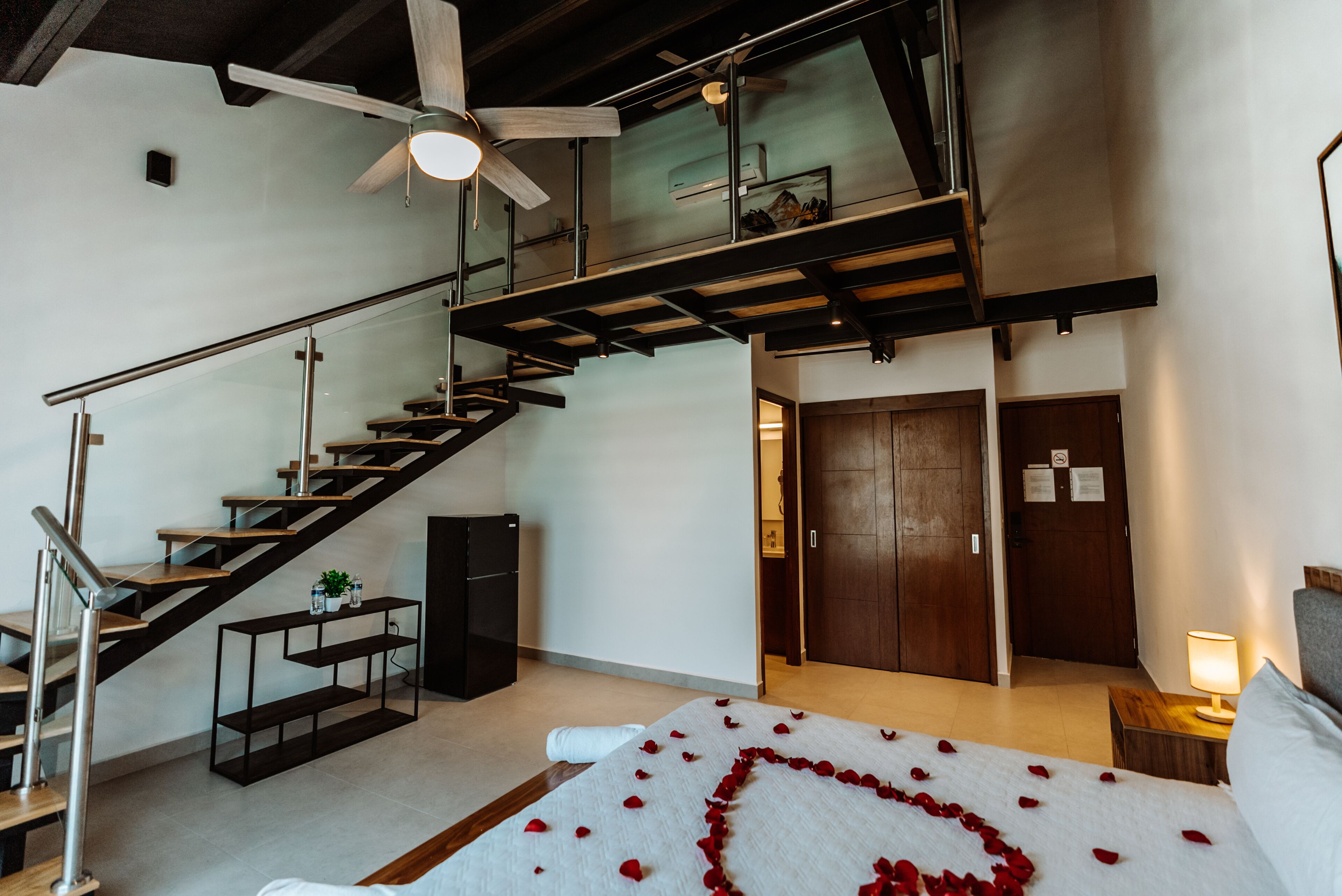 Une chambre d’hôtel moderne avec un escalier, un lit parsemé de pétales de rose, un ventilateur de plafond et une porte en bois.