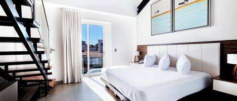 A modern hotel room with a large bed, a wooden staircase, a sliding glass door to a balcony, and two framed artworks on the wall.