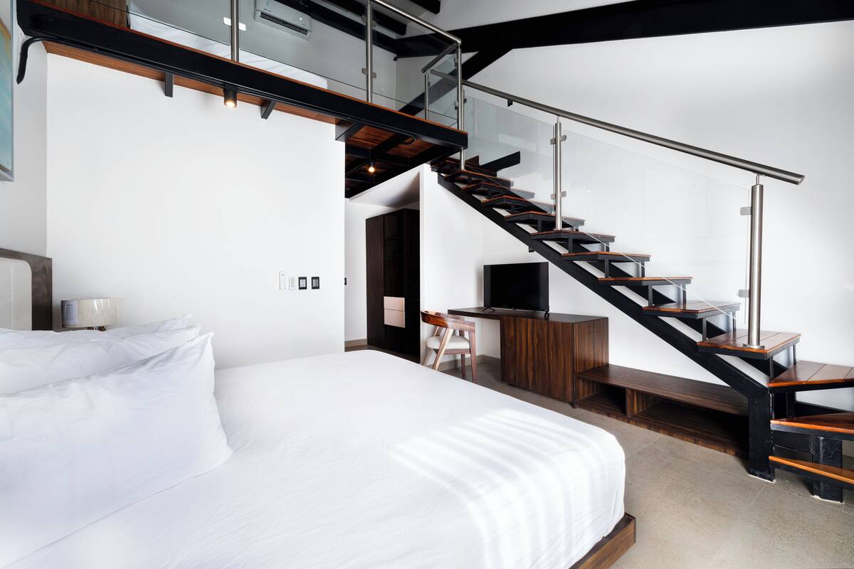 Een moderne hotelkamer met een groot bed, een bureau met stoel, een televisie en een trap met glazen balustrades.