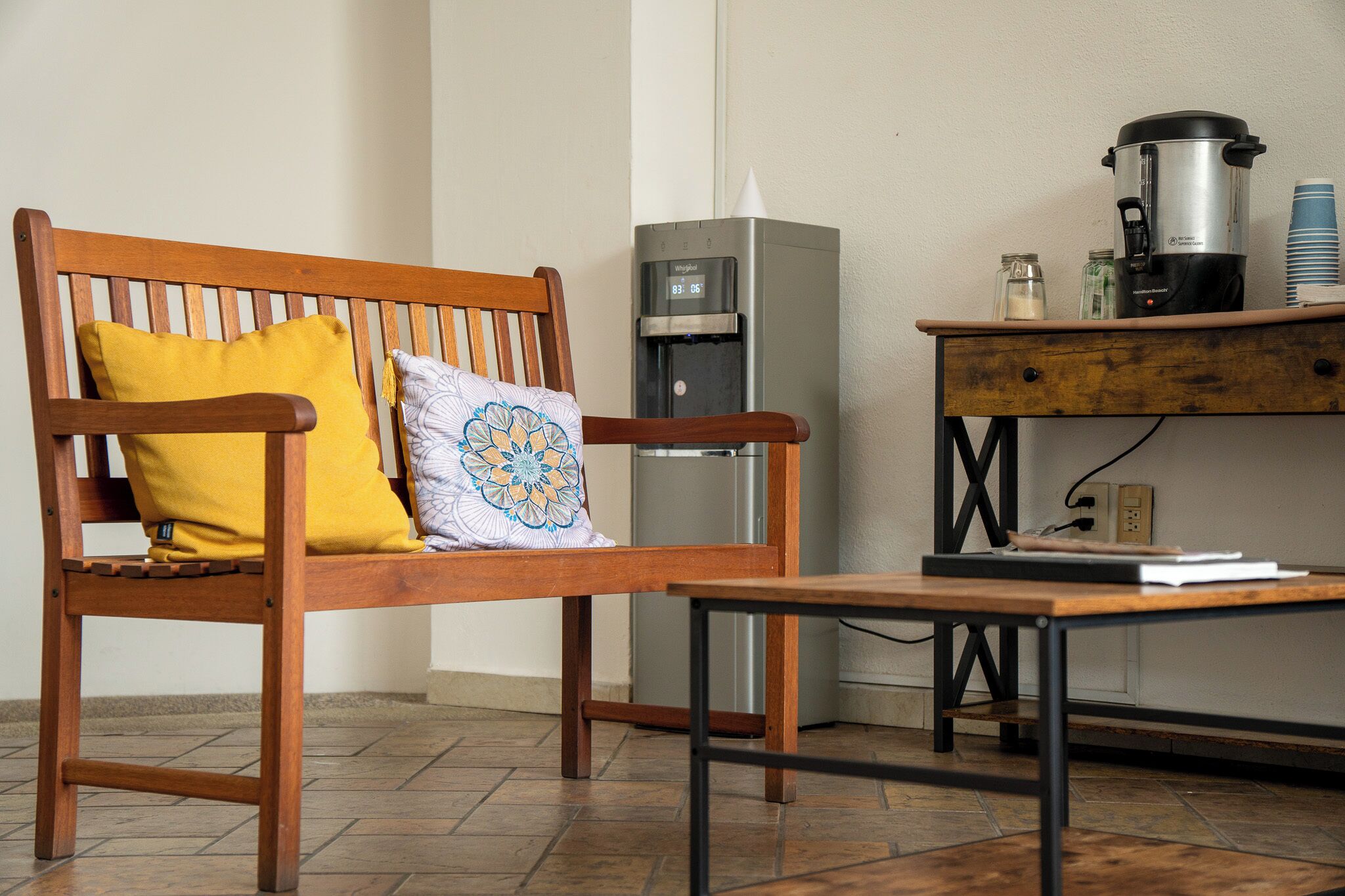 Een houten bank met een geel kussen en een decoratief kussen, een koffiezetapparaat en een kleine tafel met een boek.
