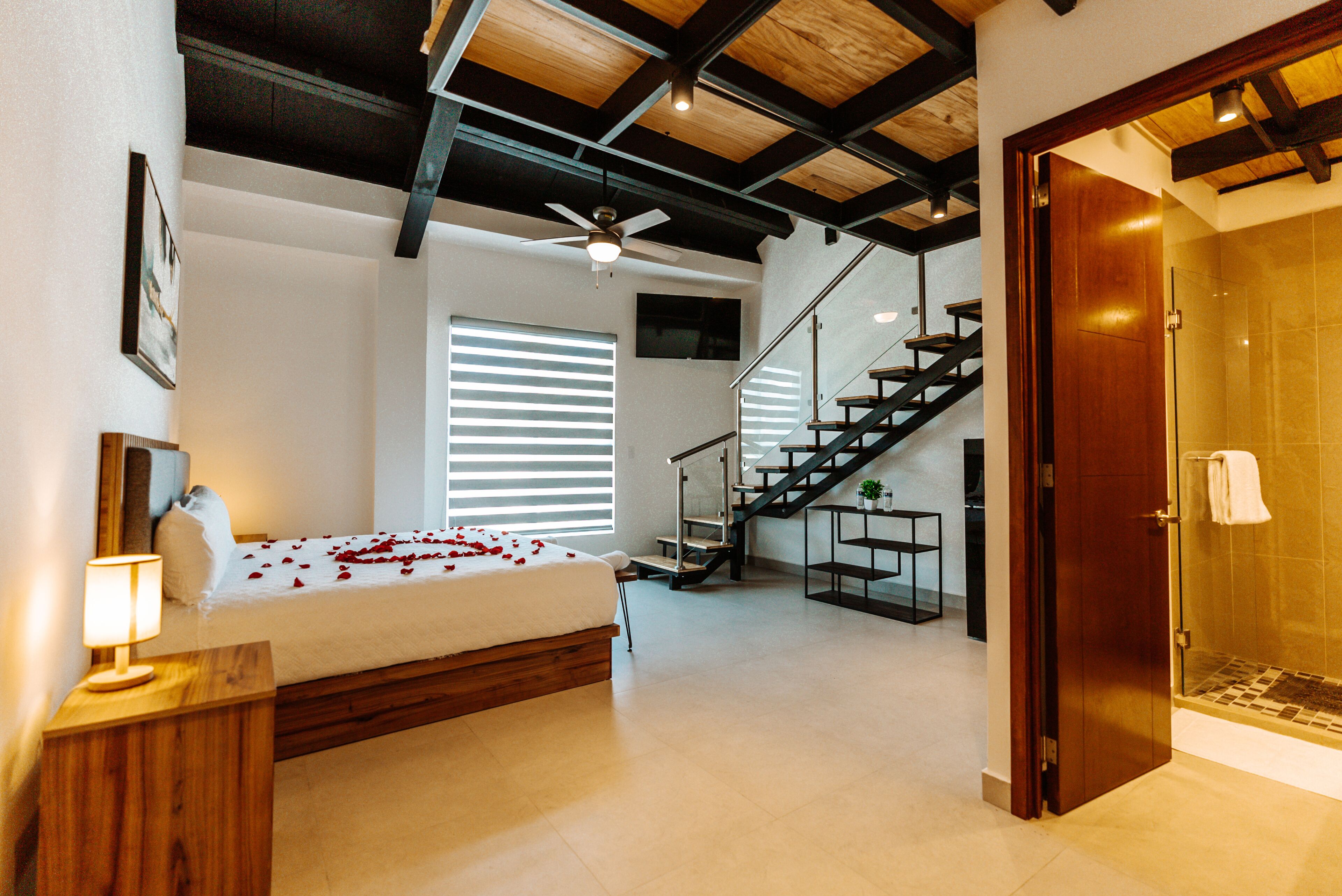 A bedroom with a bed, a bedside table, a ceiling fan, a staircase, and a bathroom with a shower.