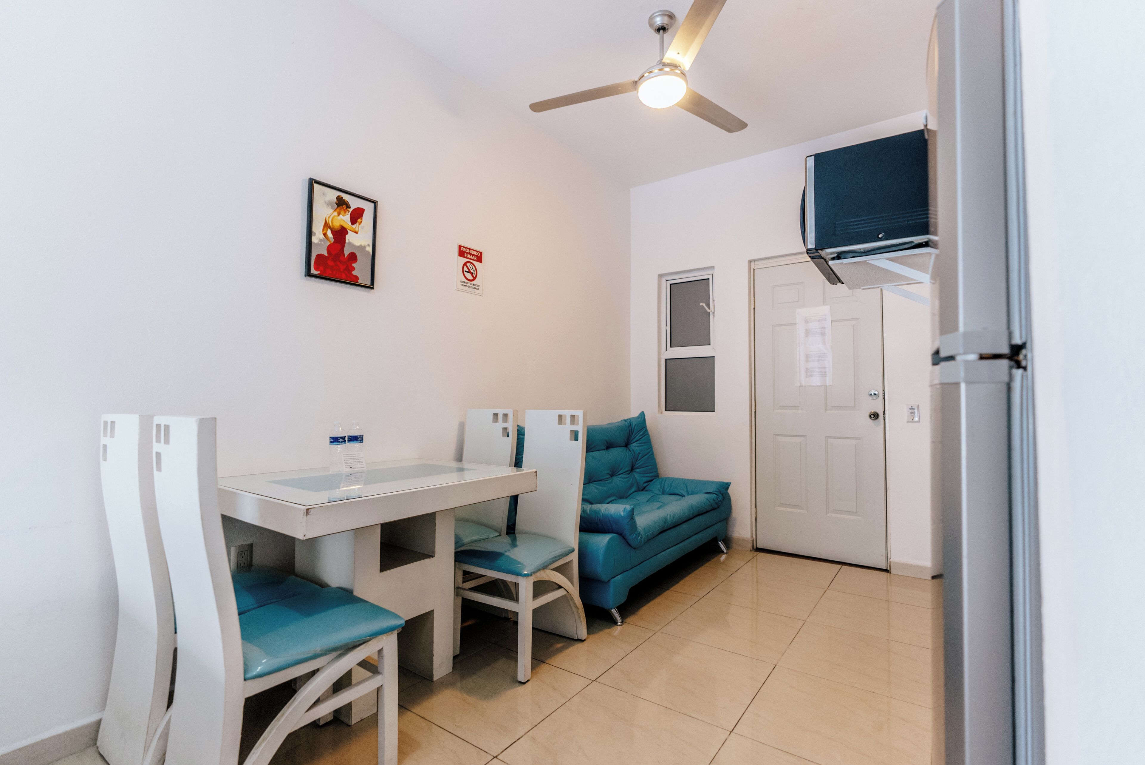 Een kleine, schone kamer met een wit bureau, twee stoelen, een blauwgroene bank, een plafondventilator en een deur met een raam.