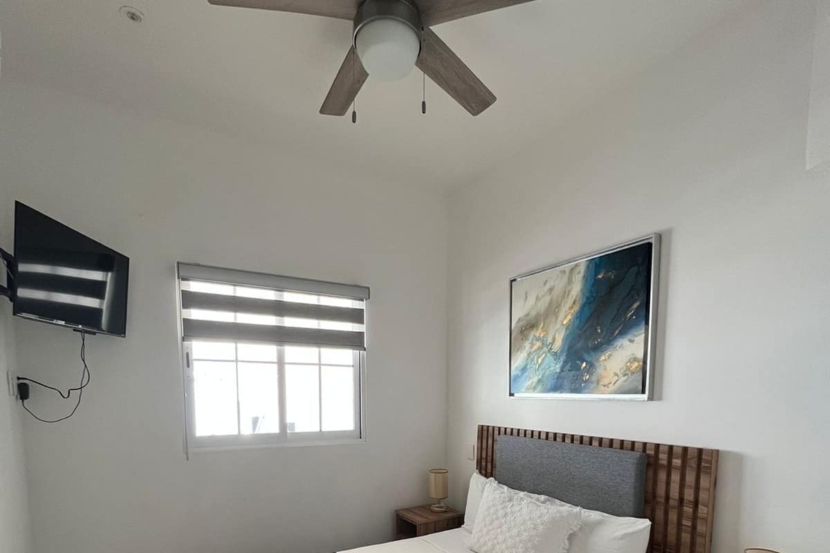 Een slaapkamer met een bed, een plafondventilator, een televisie, een raam met jaloezieën, een schilderij en een nachtkastje met een lamp.