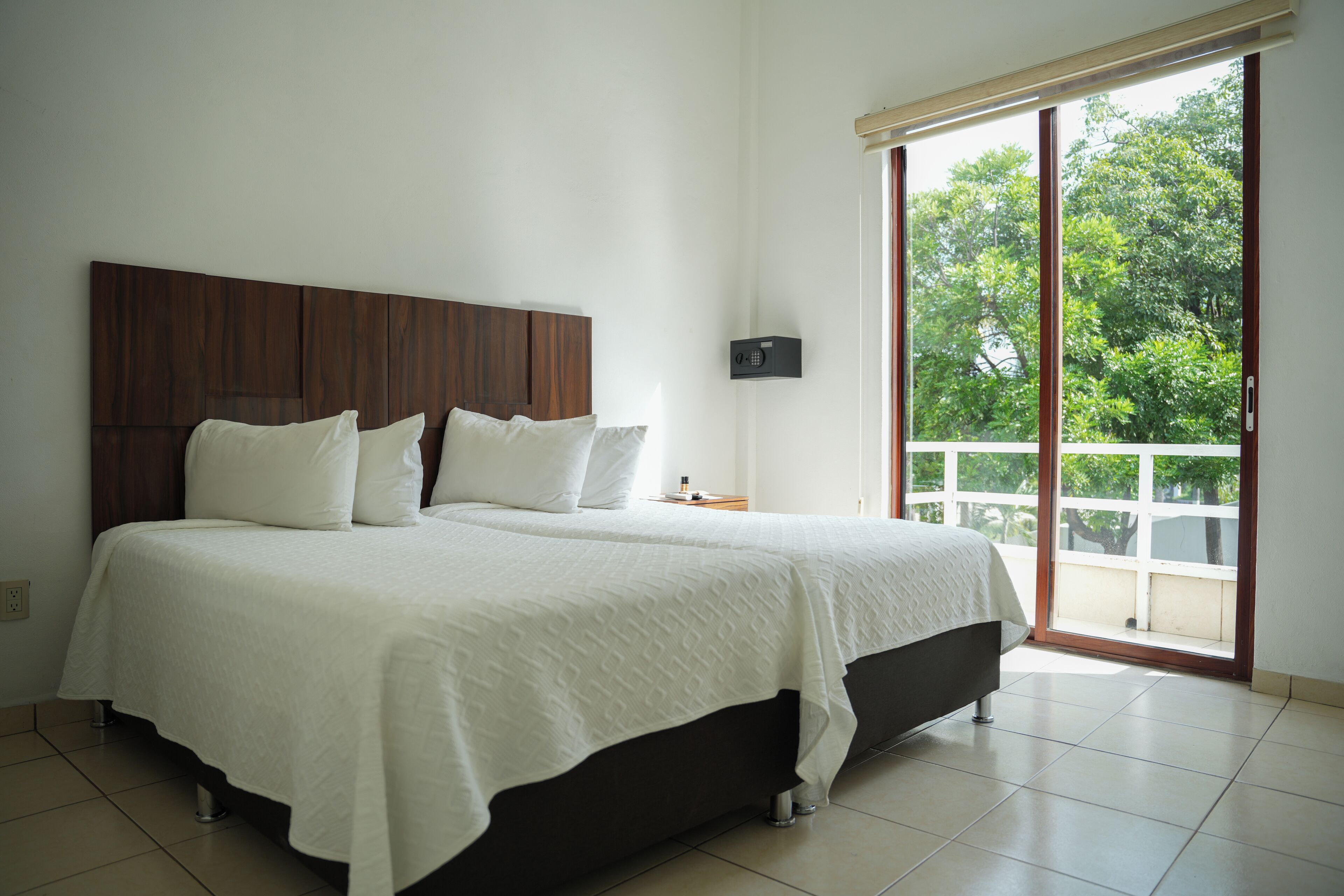 A bedroom with a large bed, a wooden headboard, a nightstand, and a view of greenery through a sliding door.