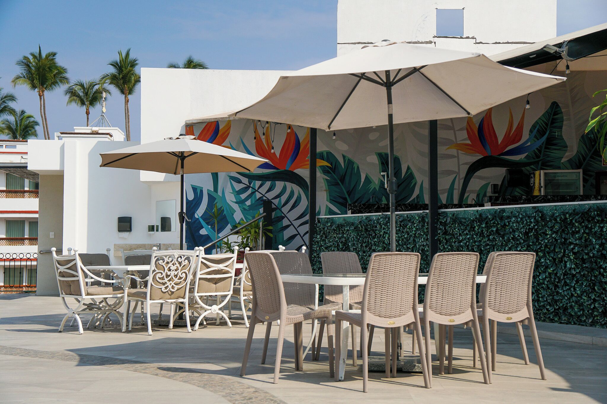 An outdoor seating area with wicker chairs and tables, umbrellas, and a mural with tropical leaves.