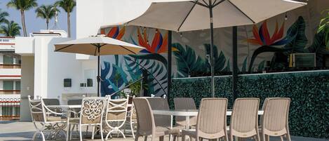 An outdoor seating area with wicker chairs and tables, umbrellas, and a mural with tropical leaves.