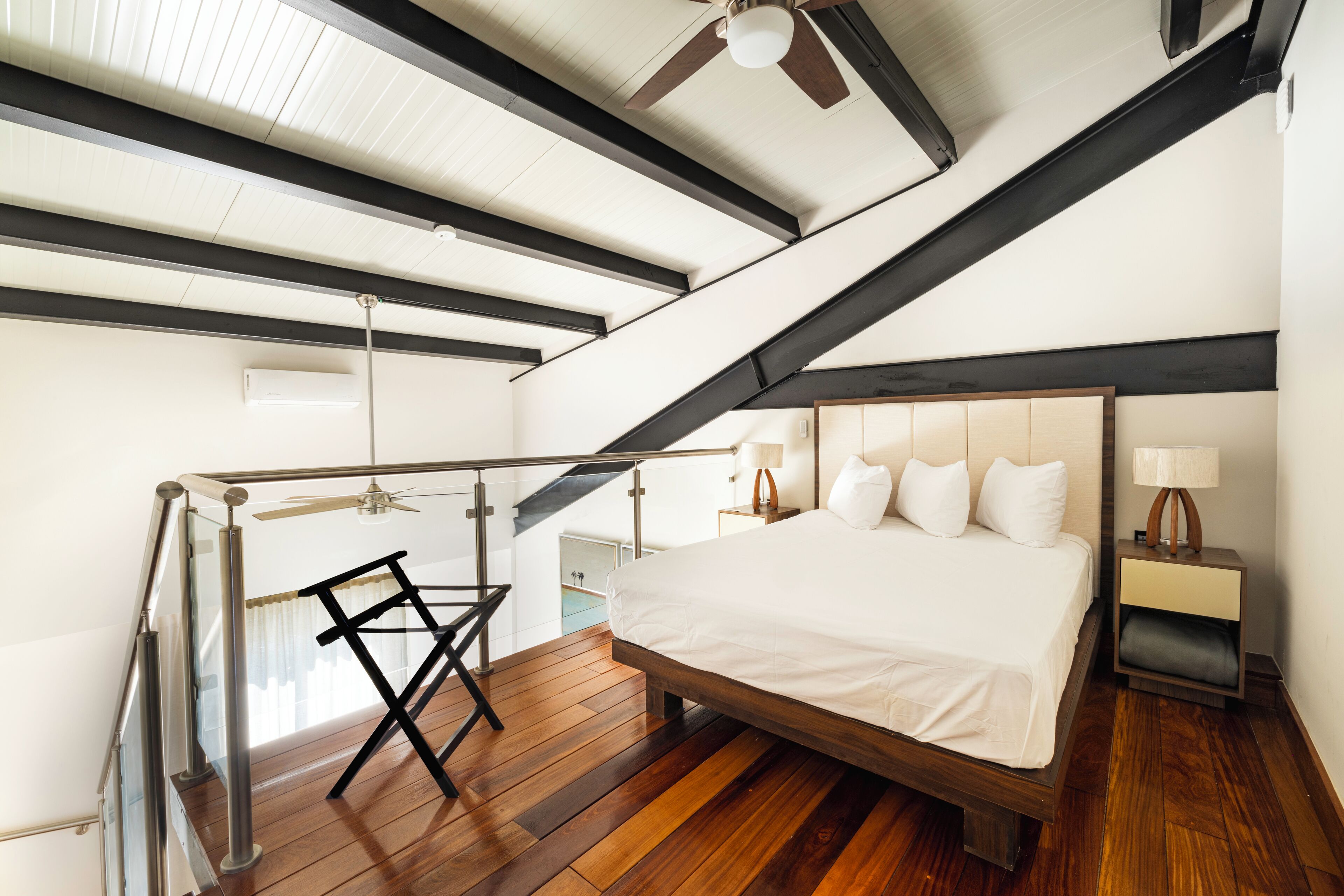 A bedroom with a wooden floor, a bed with white bedding, a nightstand, a lamp, and a ceiling fan.