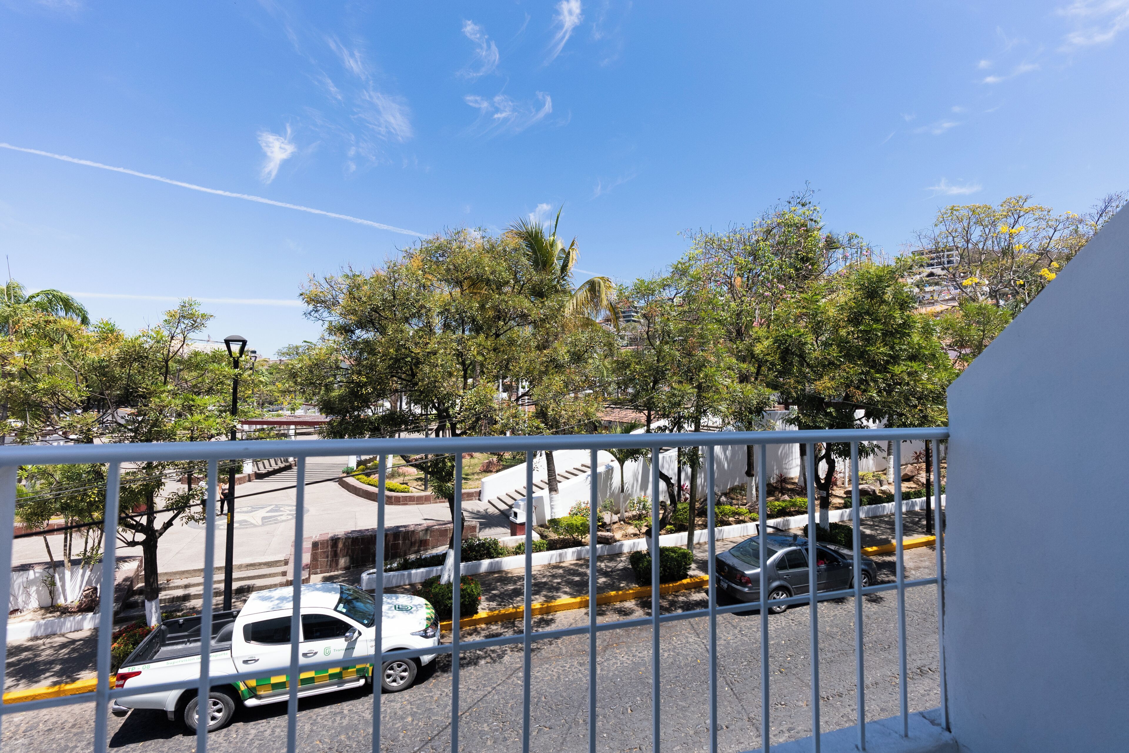 A view from a balcony with a metal railing, a street with parked cars, trees, and a clear sky.