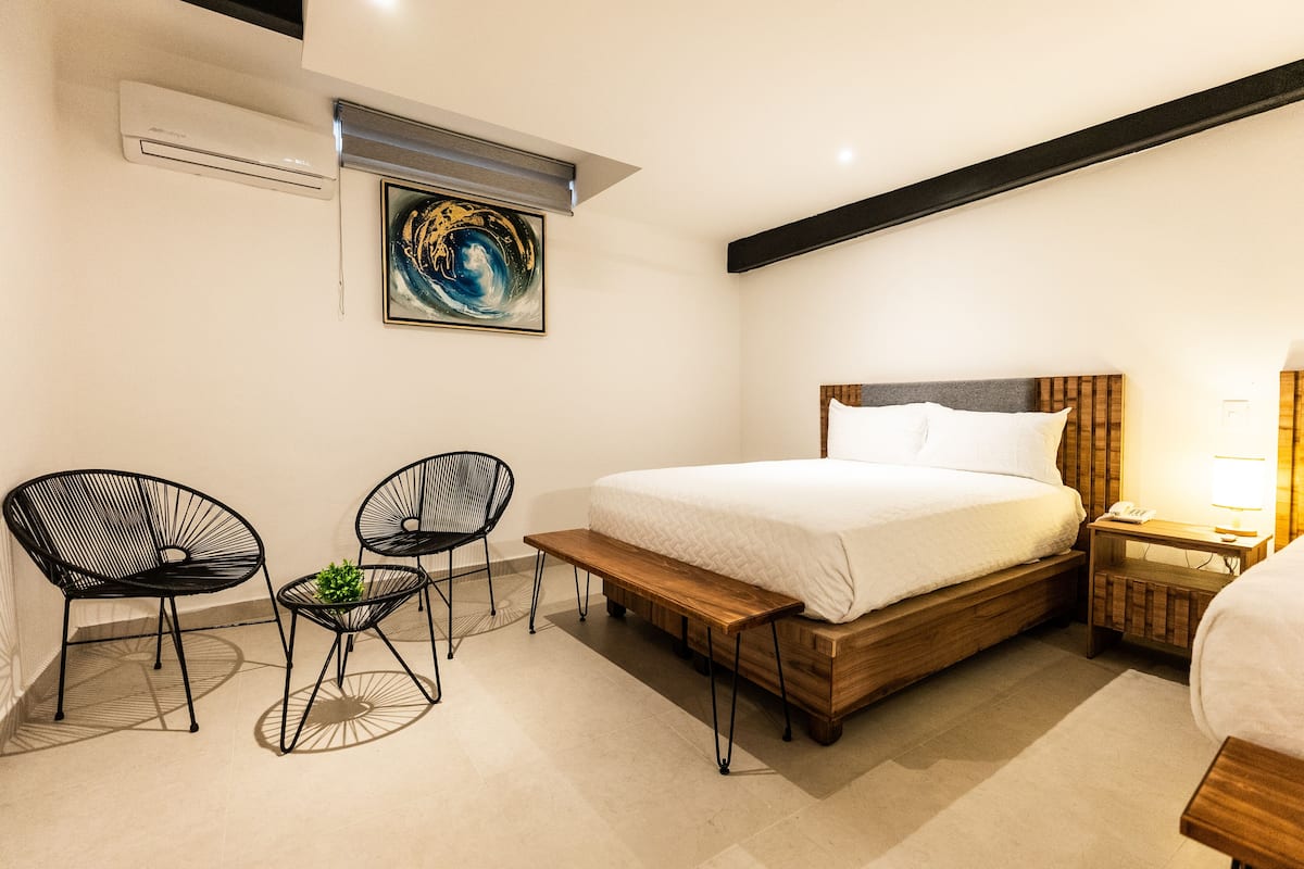 Een slaapkamer met een bed, twee stoelen, een kleine tafel, een airconditioner en een schilderij aan de muur.