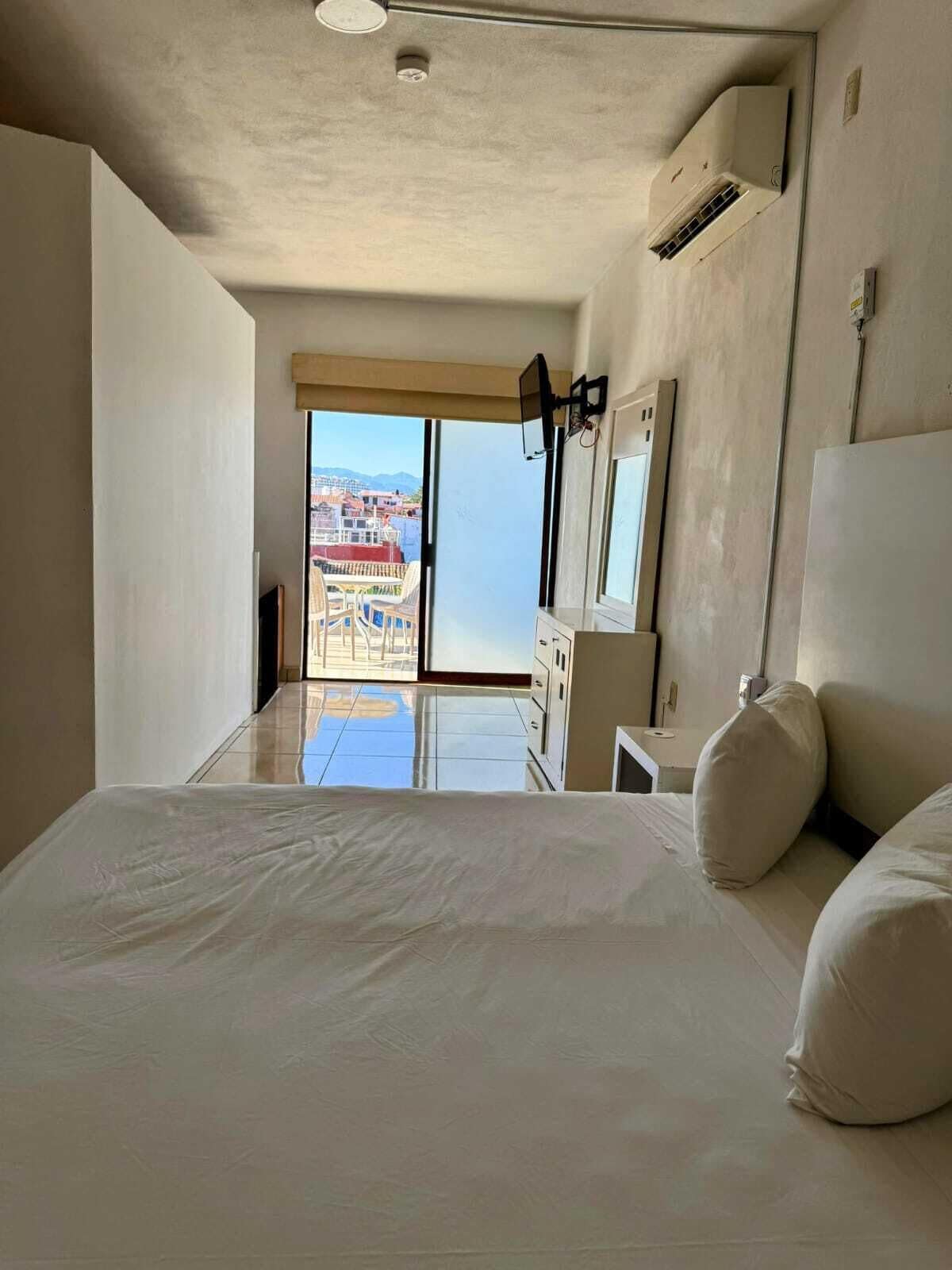 Un dormitorio con una cama grande, vistas de edificios a través de la ventana y un equipo de aire acondicionado en la pared.
