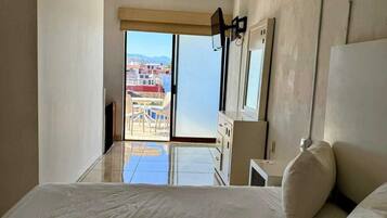 A bedroom with a large bed, a view of buildings through the window, and an air conditioning unit on the wall.